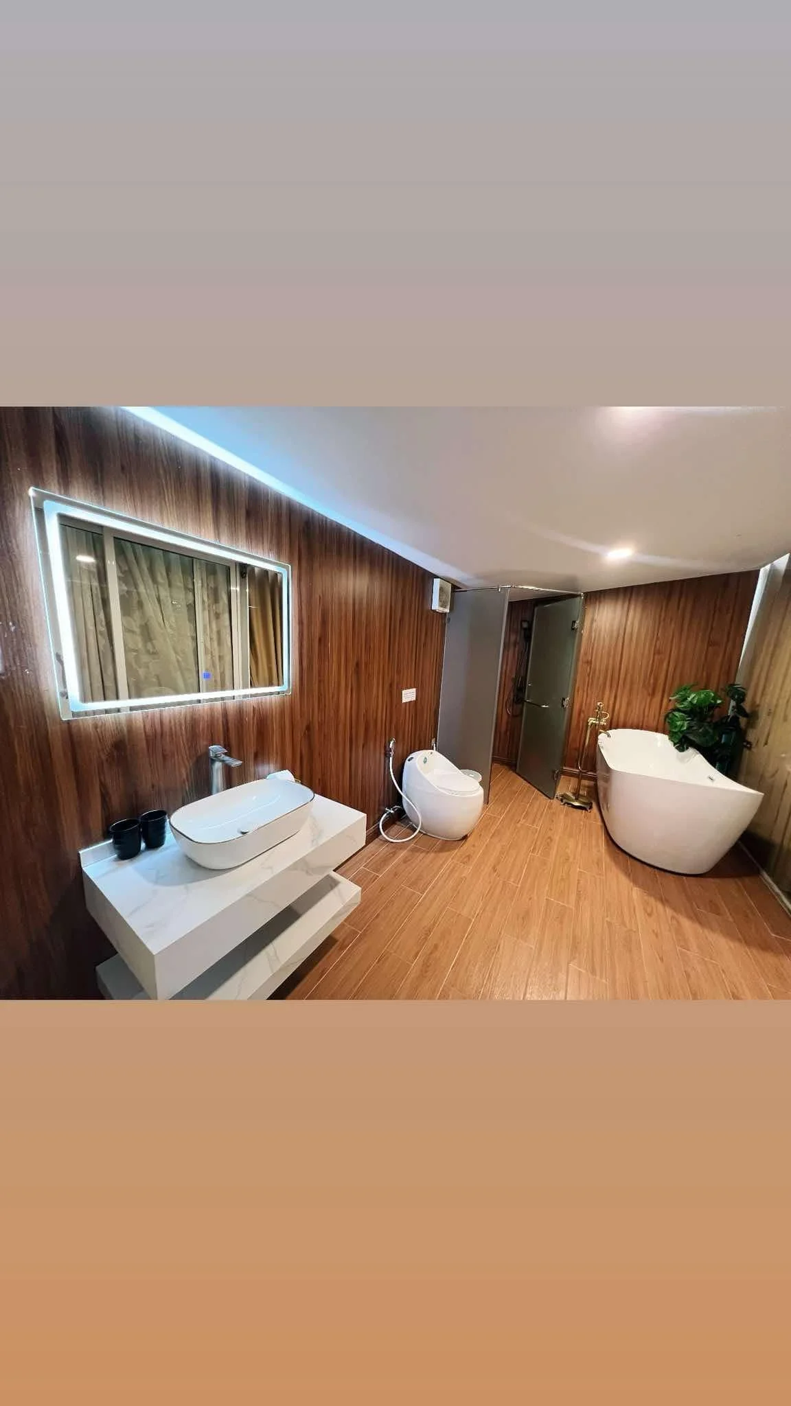 Modern bathroom with wood-panel walls, white sink on a marble countertop, a bidet, a freestanding bathtub with a plant nearby, and a mirror with LED lighting.