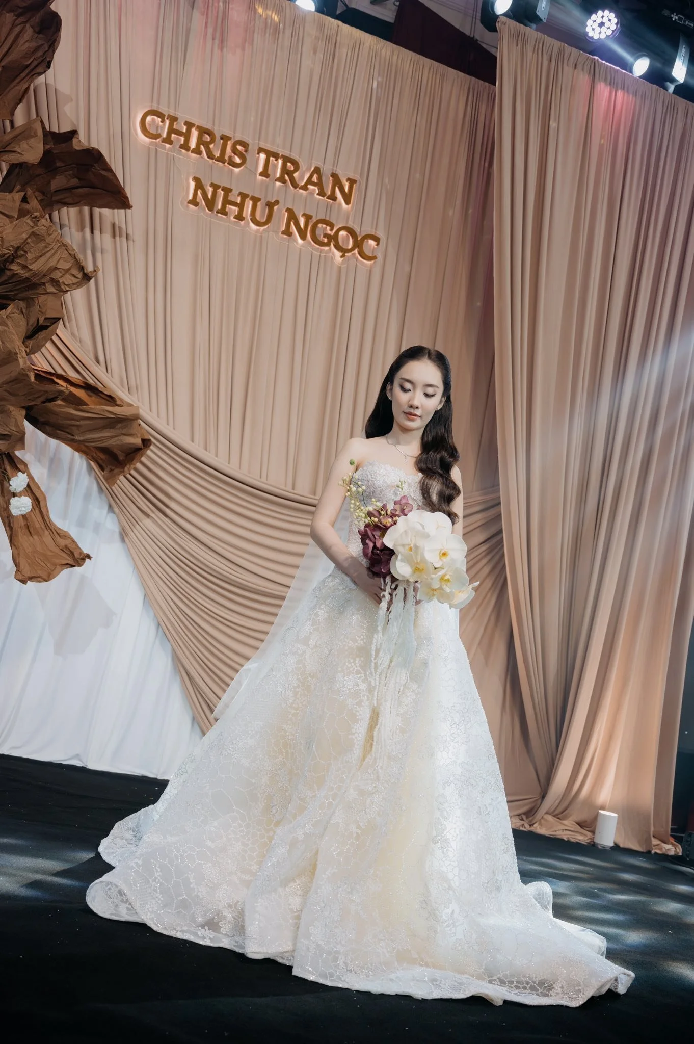 A woman in a wedding dress holding a bouquet of flowers standing in front of a curtain backdrop with the names 'Chris Tran' and 'Nhu Ngoc' illuminated above her.