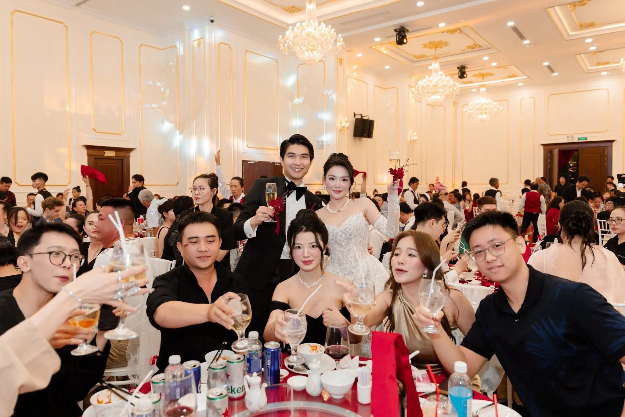 A wedding reception with the bride and groom standing together holding drinks, surrounded by seated guests at round tables in an elegant banquet hall decorated with chandeliers and gold accents.