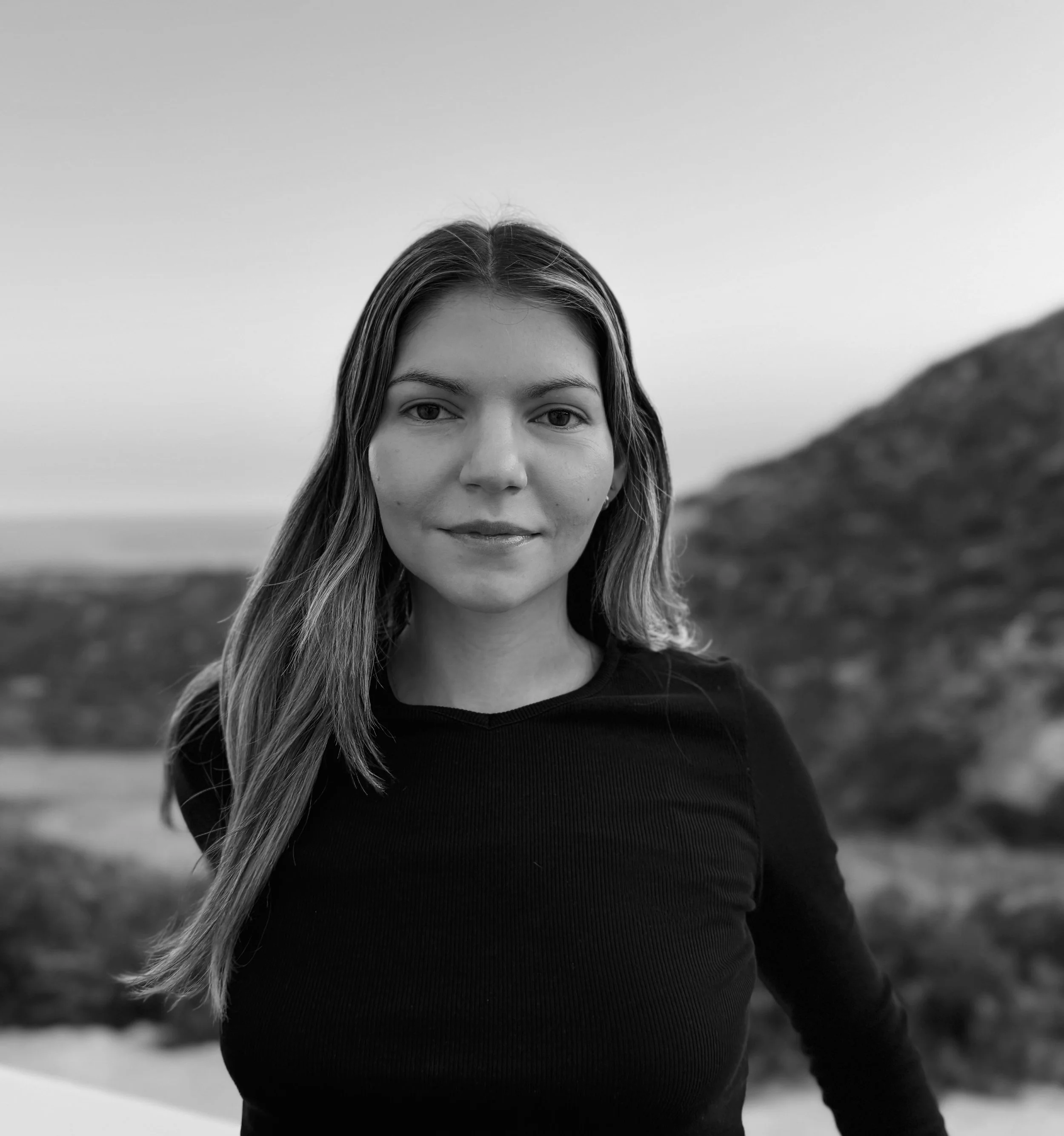 A young woman with long hair in an outdoor setting with hills or mountains in the background, smiling slightly, in black and white.