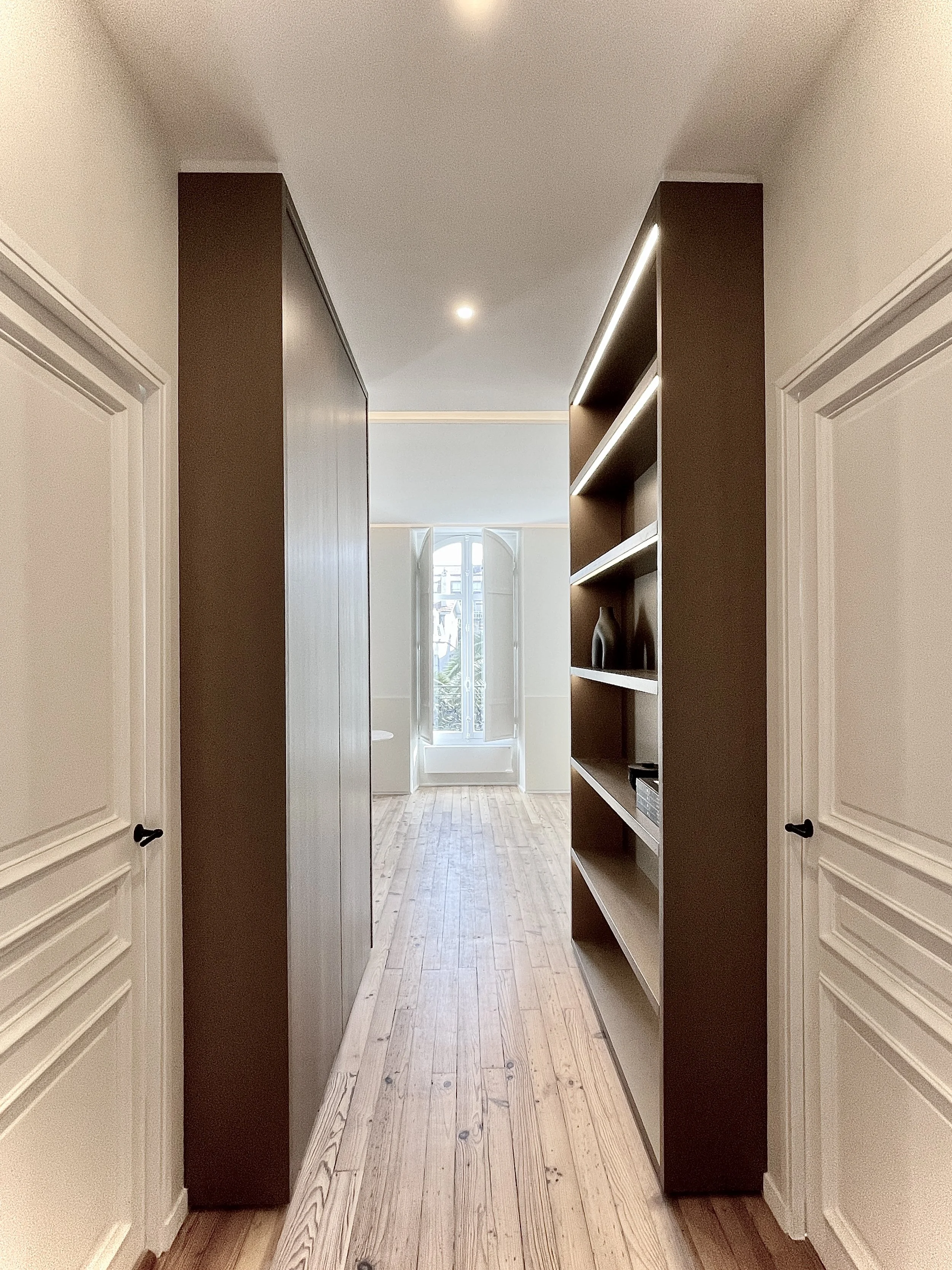 Perspective depuis l'entrée vers la zone jour de l'appartement, les deux portes mènent en zone nuit aux deux suites chambres/salle de douche, le meuble de gauche renferme une armoire buanderie et penderie, celui de droite une large bibliothèque.    