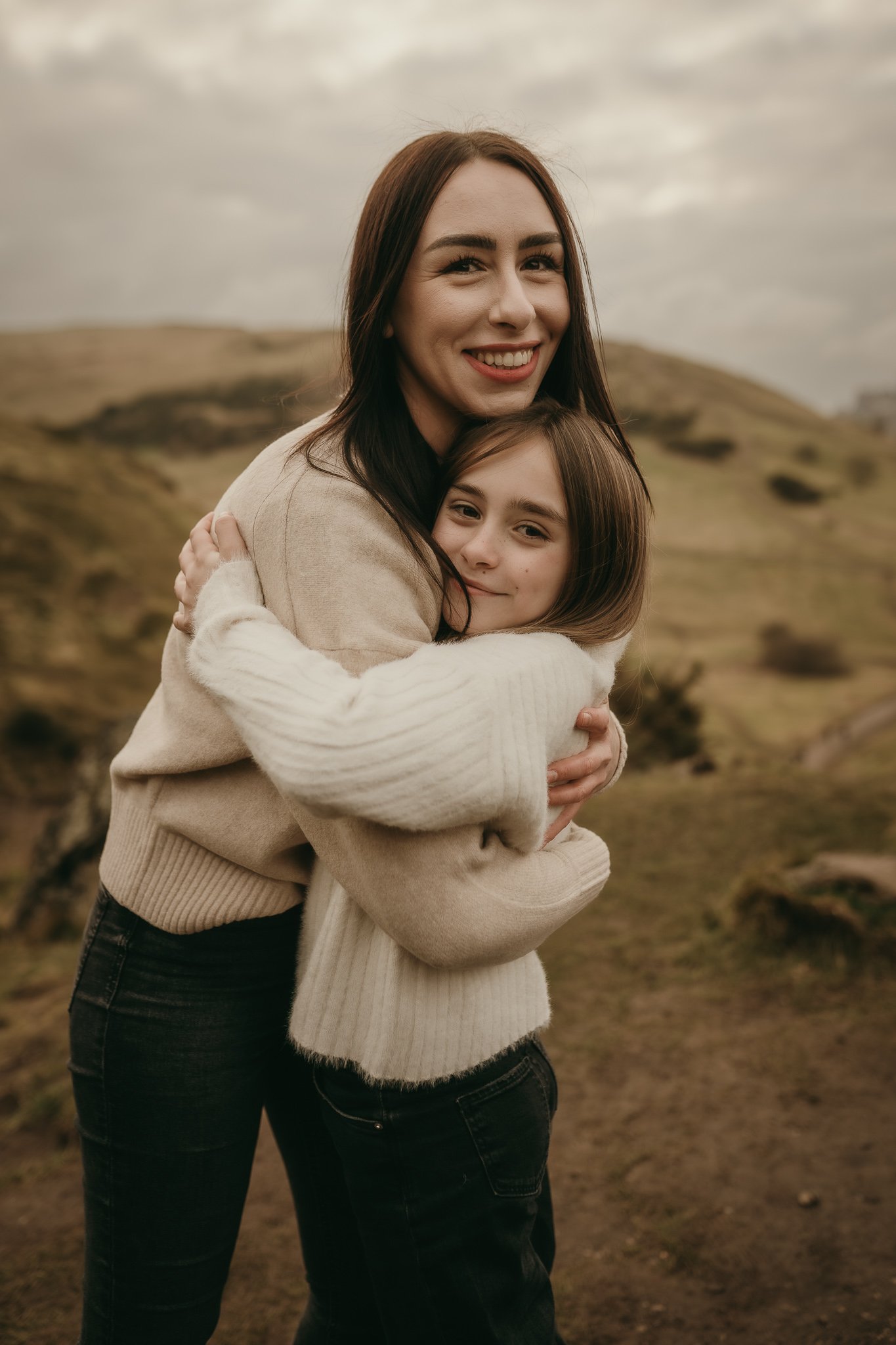 A woman and a child hugging outdoors in a hilly landscape, both smiling and wearing sweaters.