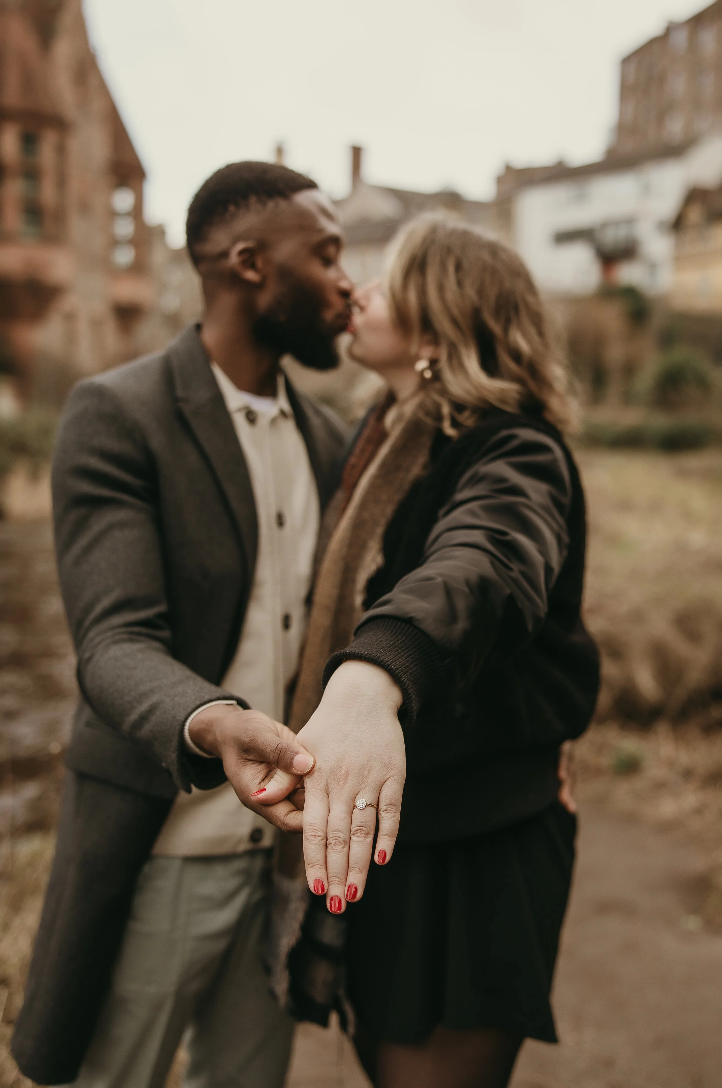 Close-up of engagement ring with a kissing couple in the background, natural proposal photography in Edinburgh.