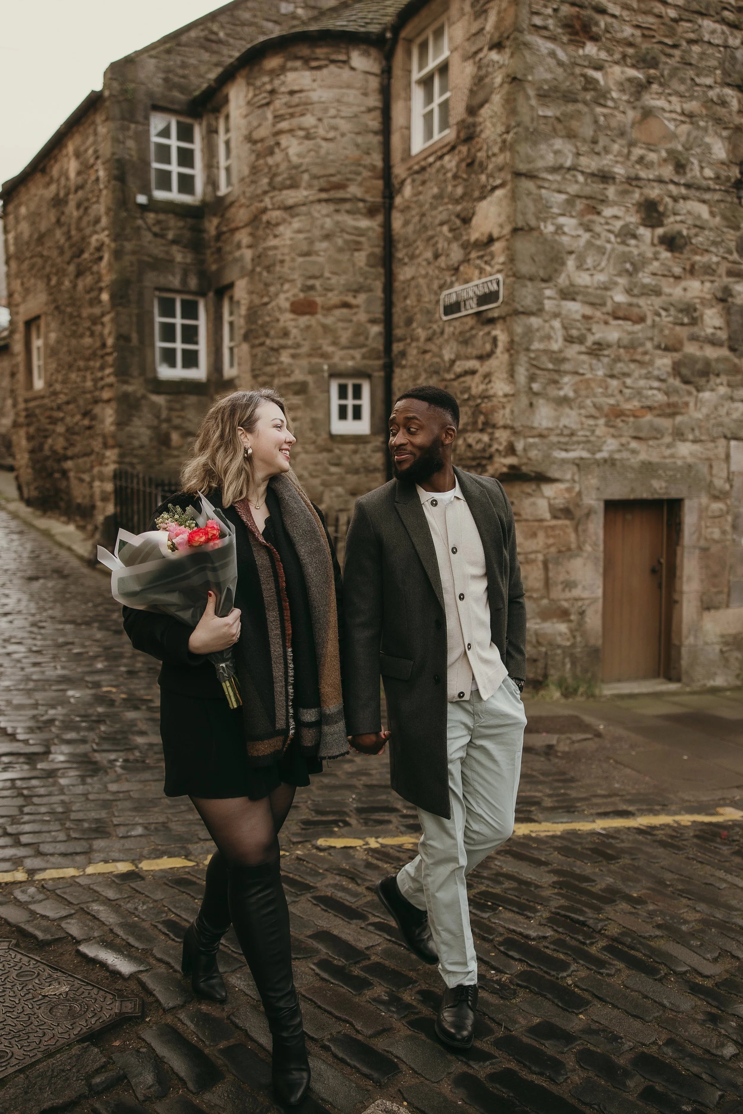 Newly engaged couple walking hand in hand through the historic streets of Edinburgh with flowers , natural and relaxed proposal photos.