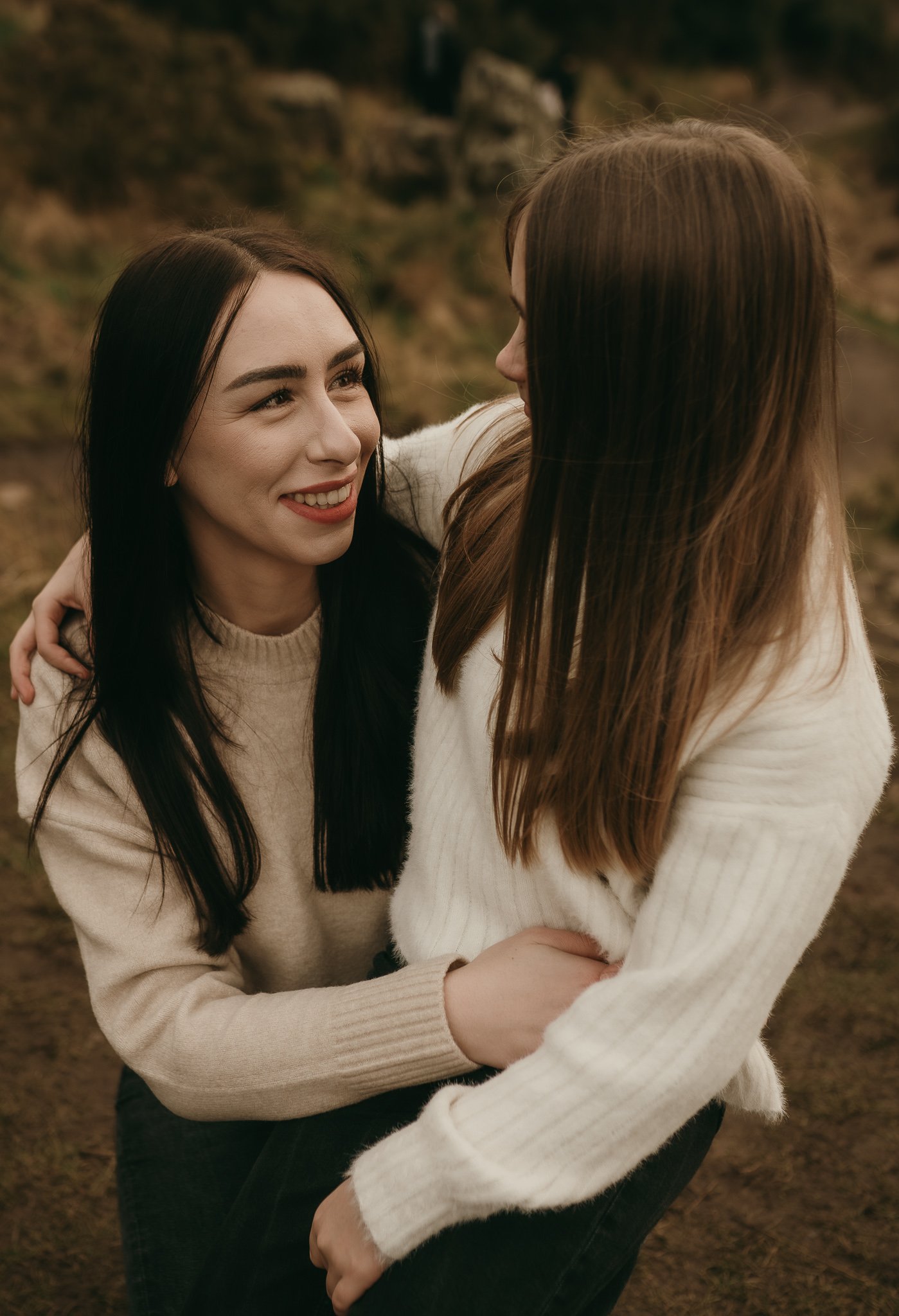 Two people in sweaters smiling at each other outdoors, one person embracing the other.