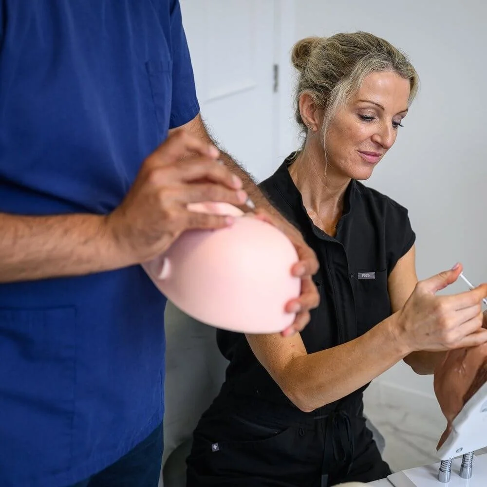 A woman with gray hair in a black shirt holding an injection syringe, a person wearing blue scrubs holding a pink breast model, in a clinical setting.