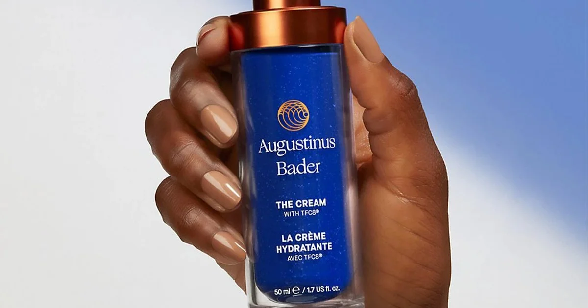 A hand holding the blue glass bottle of Augustinus Bader The Cream.