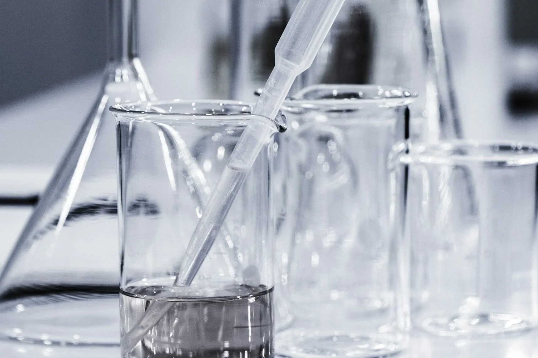 Laboratory glassware on a bench: beakers, a flask and a pipette resting in a measuring cylinder, in cool grey light.
