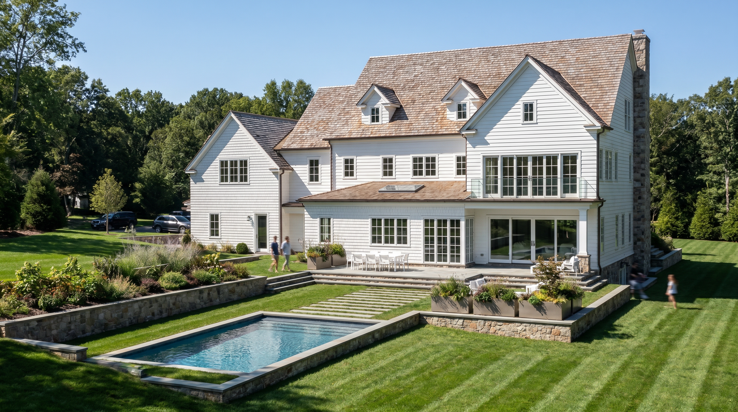 Large white house with multiple gabled roofs and a stone chimney, surrounded by a landscaped garden, swimming pool, and outdoor patio area with people walking and sitting, with tall trees in the background.