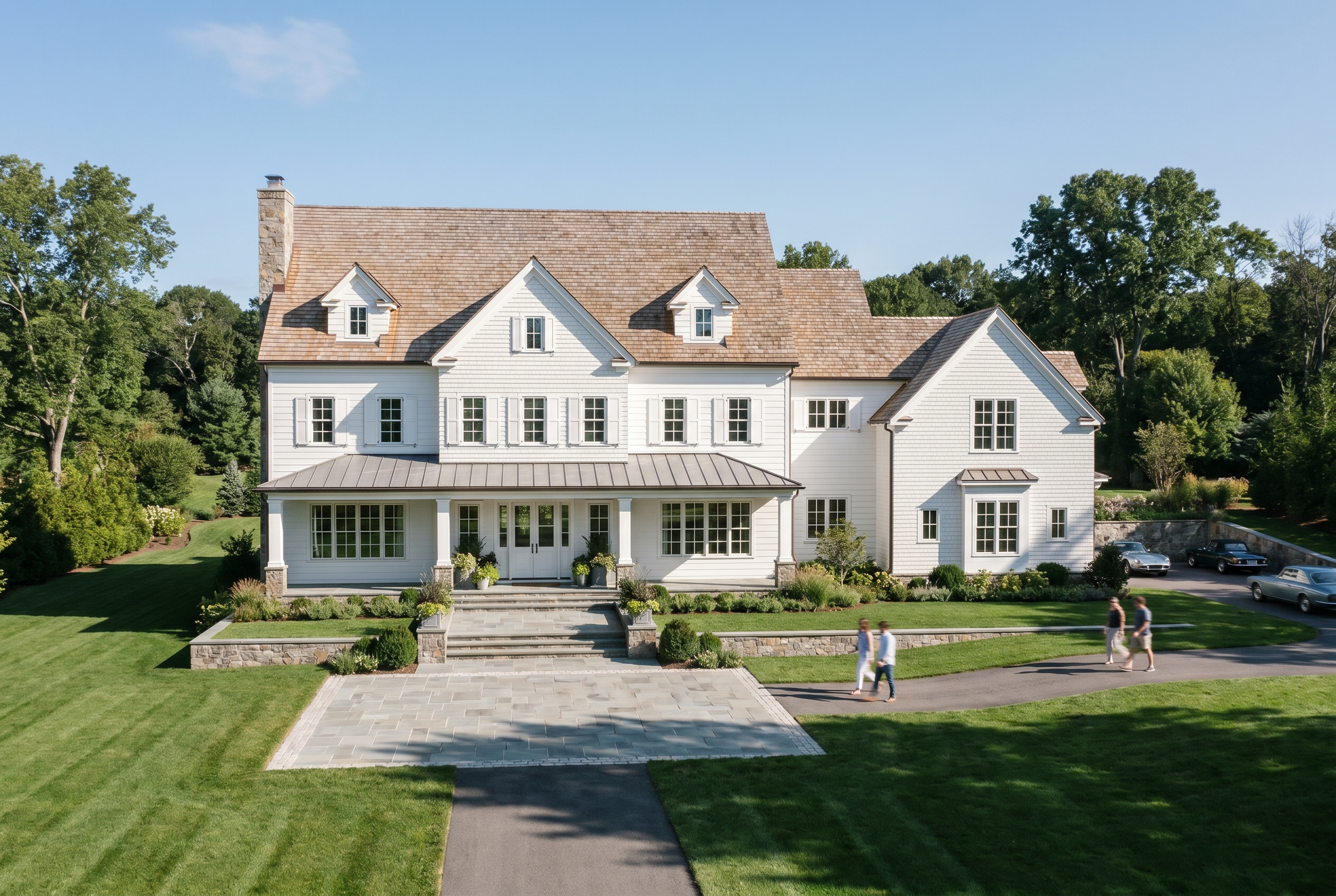 A large white house with multiple stories, gabled roofs, and numerous windows, set in a well-manicured lawn with a stone pathway, surrounded by trees on a sunny day.