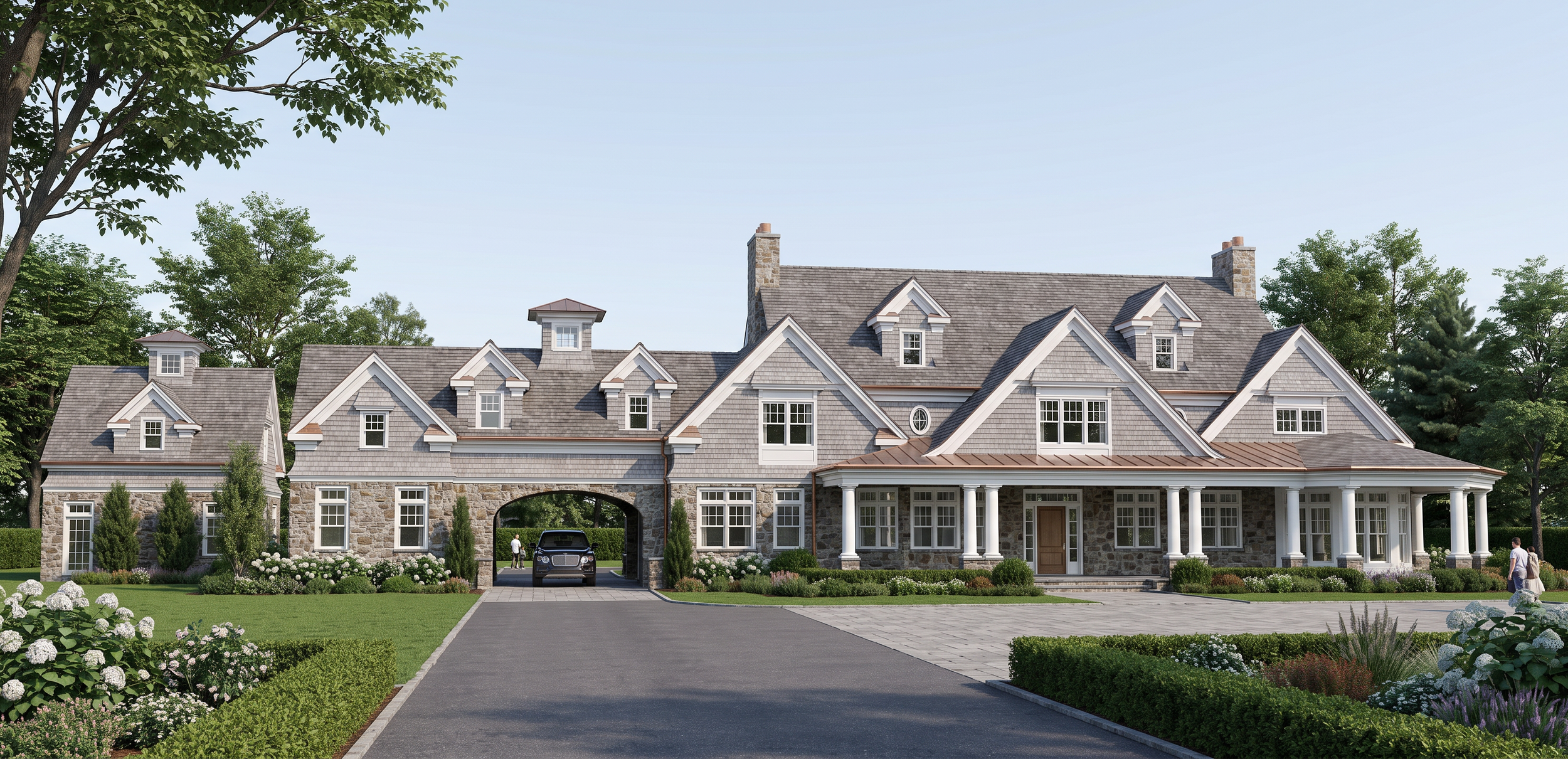 A large, elegant house with stone and gray siding exterior, multiple gables, a front porch with white columns, and surrounding landscaped garden with green trees, bushes, and flowers.