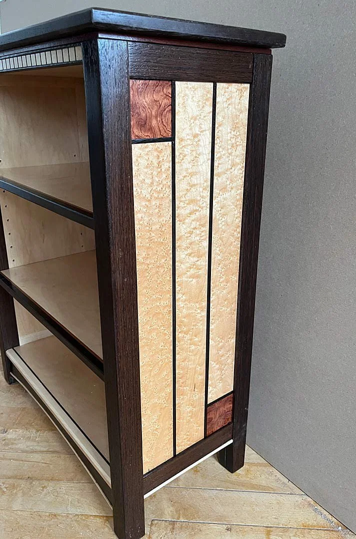 Wooden cabinet with open shelves and decorative paneling on the side, featuring light and dark wood tones, standing on a wooden floor.