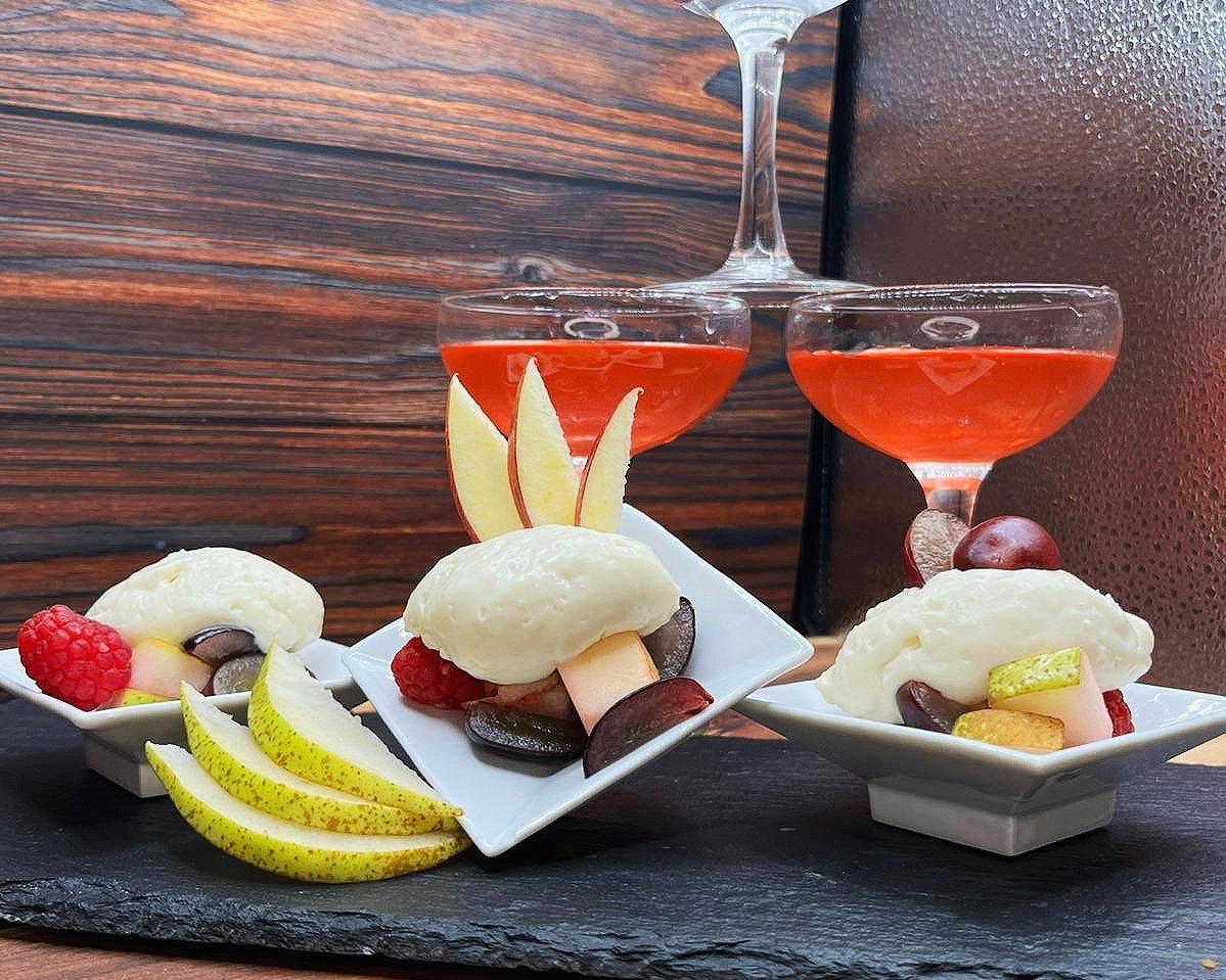 Dessert in kleinen Schalen mit Früchten wie Birnen, Äpfeln, Himbeeren und Trauben, garniert mit weißer Creme. Im Hintergrund zwei Gläser mit rötlichem Getränk auf dunklem Untergrund.