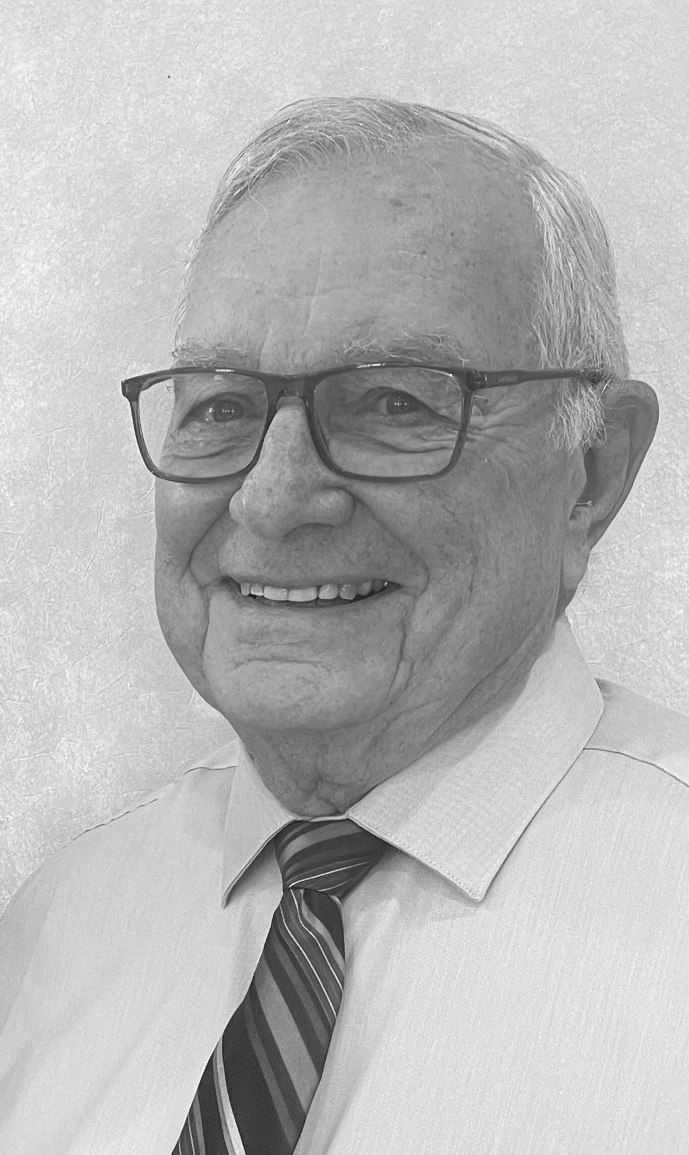 Black and white portrait of an elderly man wearing glasses, a light-colored shirt, and a striped tie, smiling.