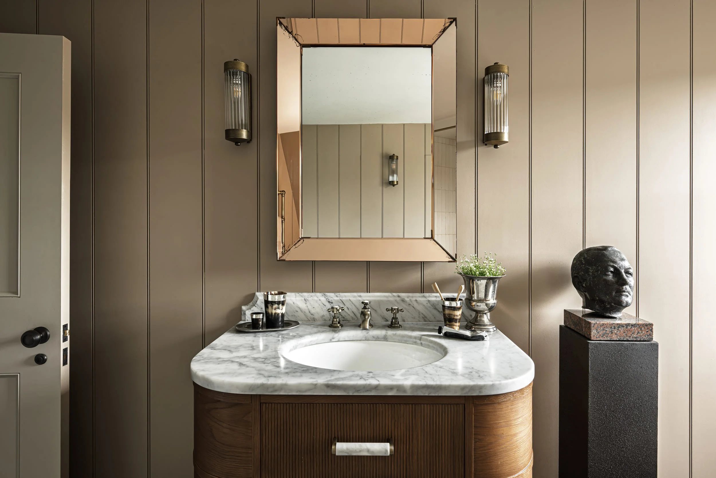 A bathroom interior design by Atelier Otis featuring a large rectangular mirror, wall-mounted lights, a potted plant, decorative cups, and a bust sculpture on a pedestal. Atelier Otis, an interior design studio based in Bath working across the globe.
