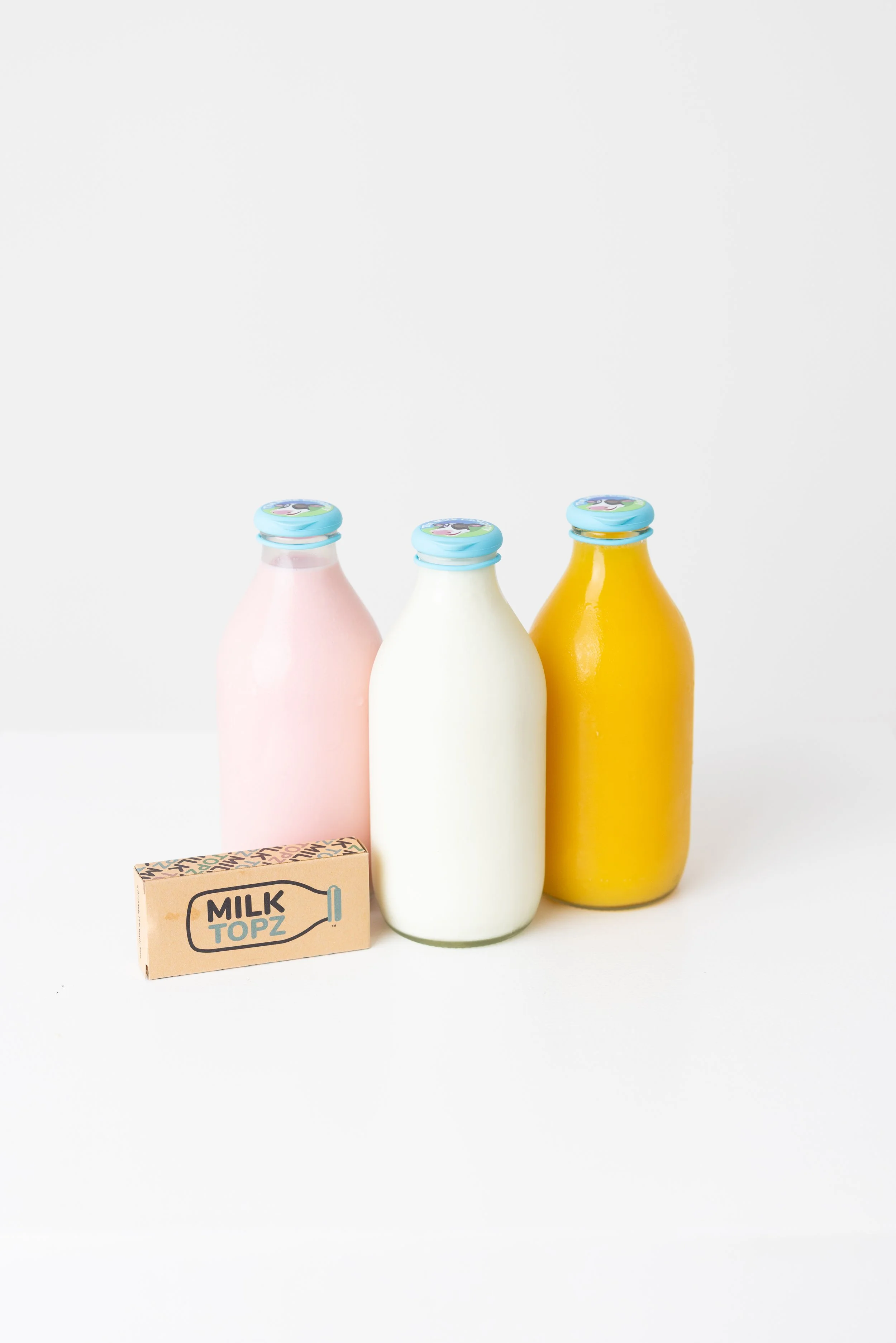 Three glass bottles with pastel-colored liquid, pink, white, and yellow, with blue caps, and a small cardboard box labeled 'Milk Topz' in front of them, all against a white background.