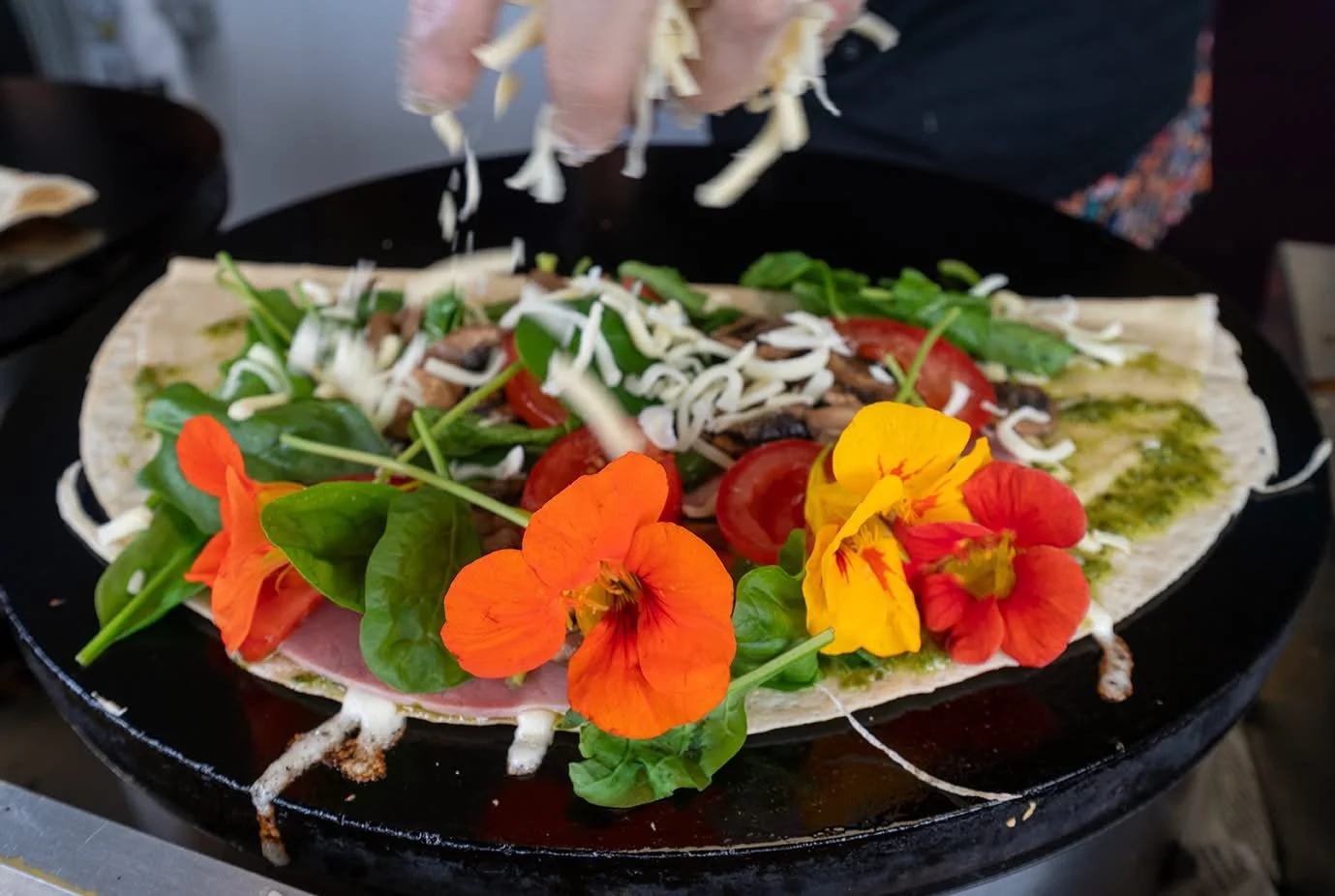 Fresh savory crêpe garnished with rocket, spinach, cheese, and edible orange flowers at a street food market.