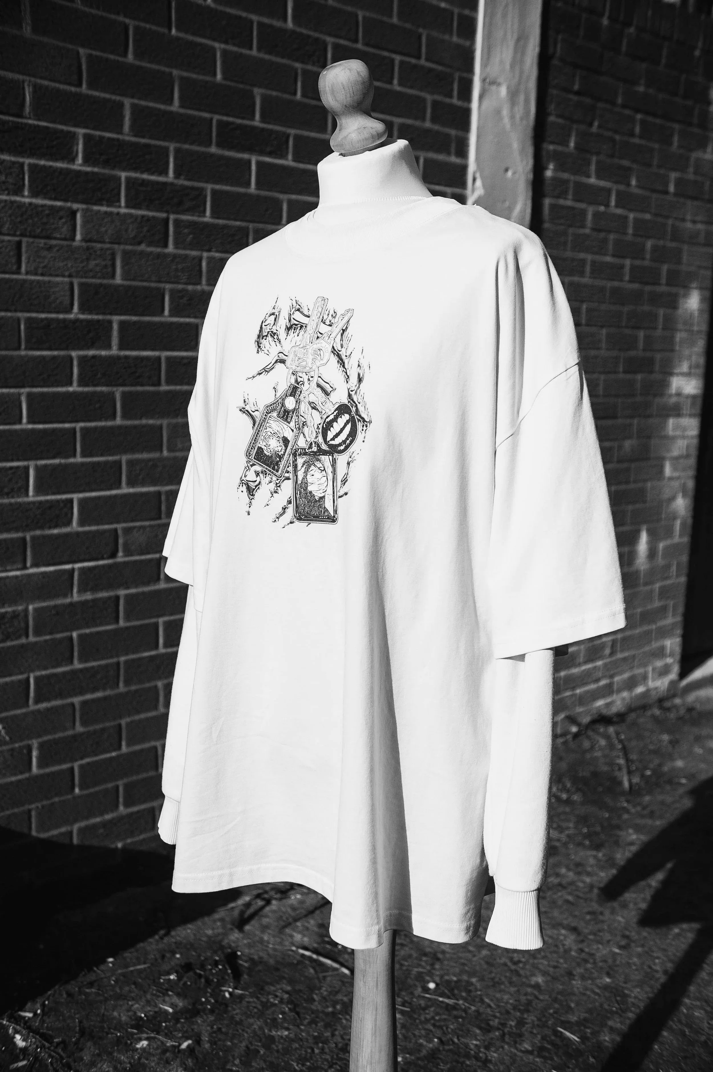 Black-and-white photo of a wooden mannequin wearing an oversized white graphic T-shirt against a brick wall.