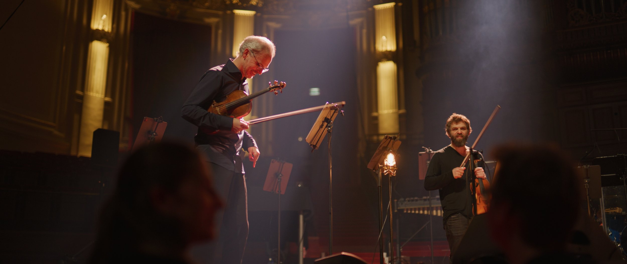 Two violinists performing on stage with music stands and warm lighting