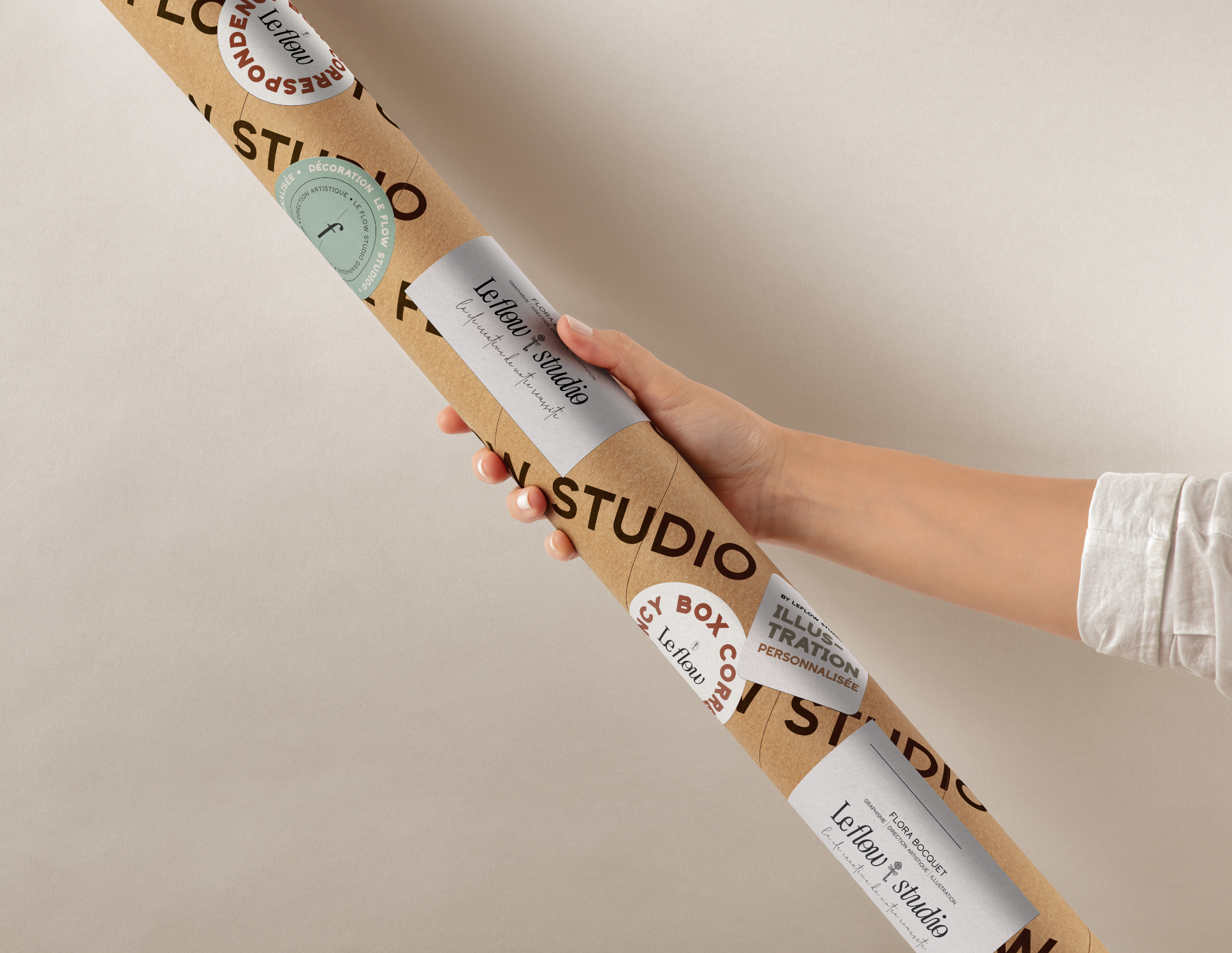 Main tenant un tube en carton marqué "Le Flow Studio", utilisé pour la livraison de décorations murales ou œuvres d'art. Plusieurs autocollants, dont certains mentionnent "illustration personnalisée", sont visibles sur le tube.