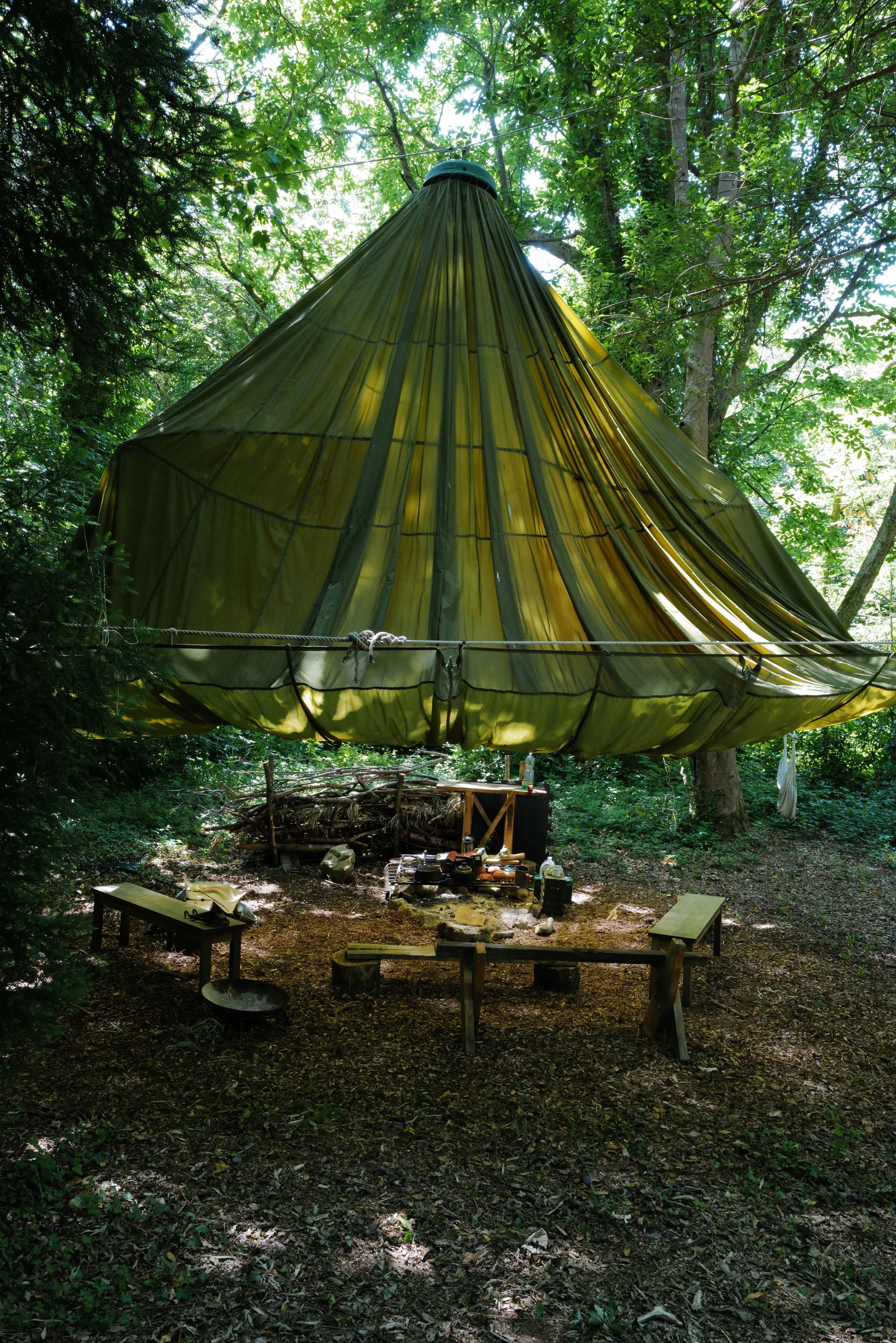 A large, rounded, yellow-green tent suspended and hanging in a forest surrounded by trees and greenery, with a small outdoor workspace beneath it.