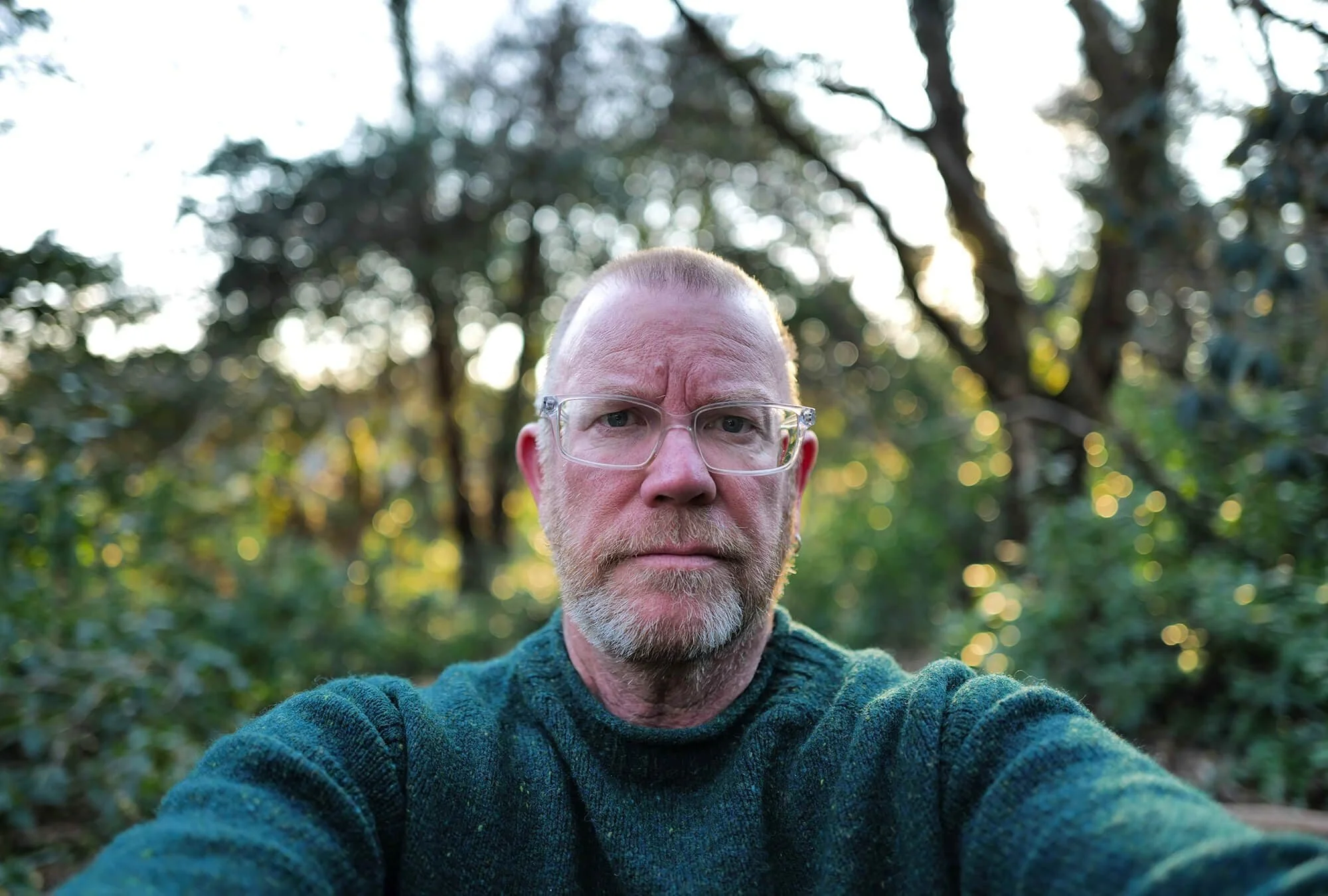 A man with glasses and a beard taking a selfie outdoors with trees and sunlight in the background.