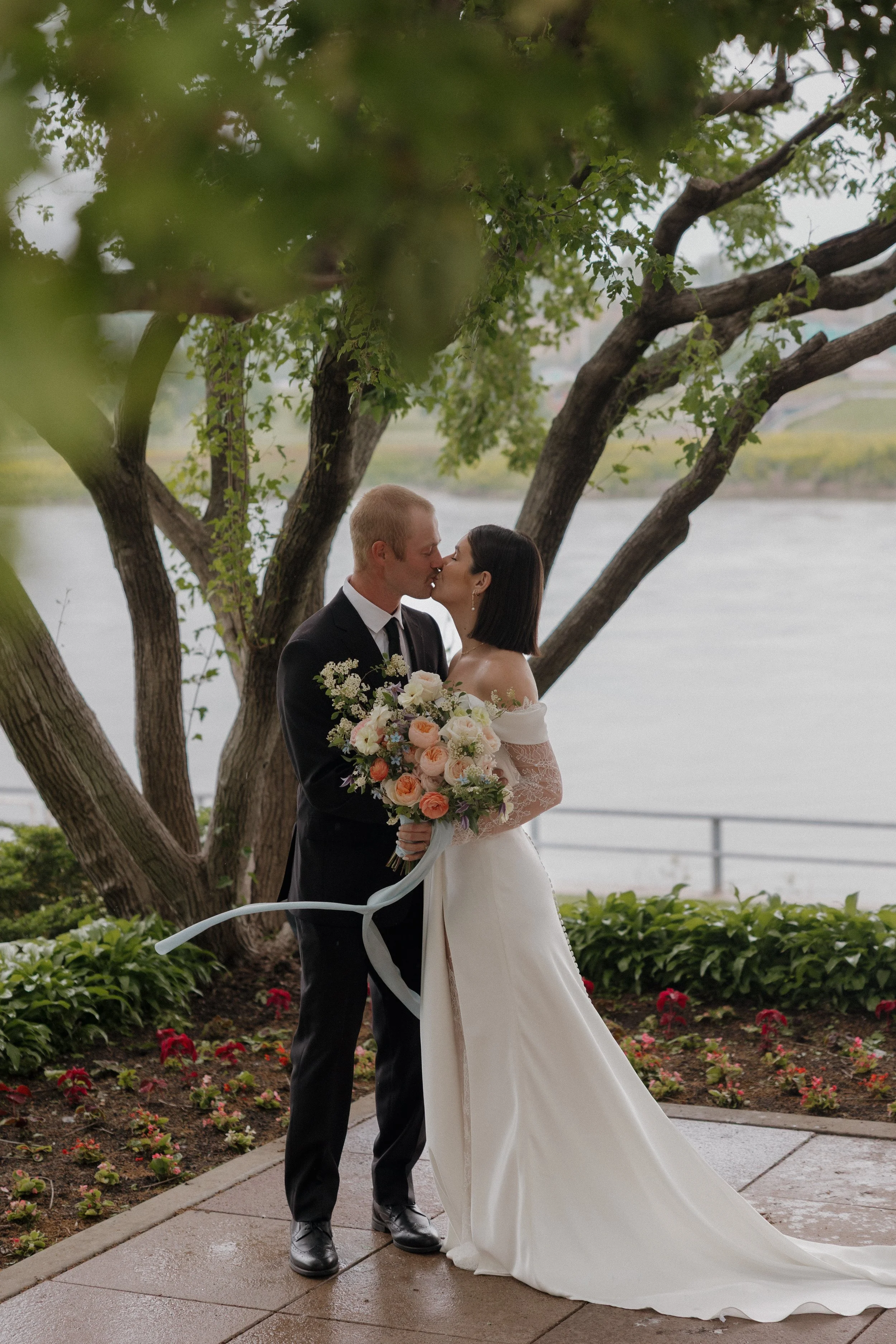 A woman in a white dress holds a large bouquet of mixed flowers including dahlias, cosmos, and other blooms with green foliage, against a plain light background.