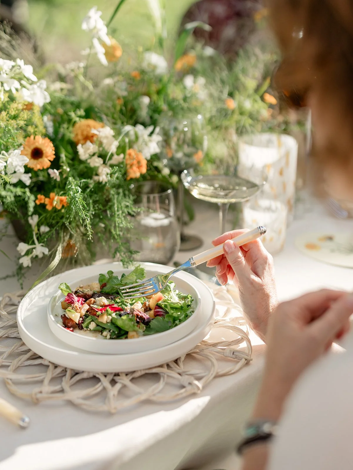 A person is eating a salad at a table decorated with a large floral arrangement with white and orange flowers, glasses of white wine, and a wicker placemat.