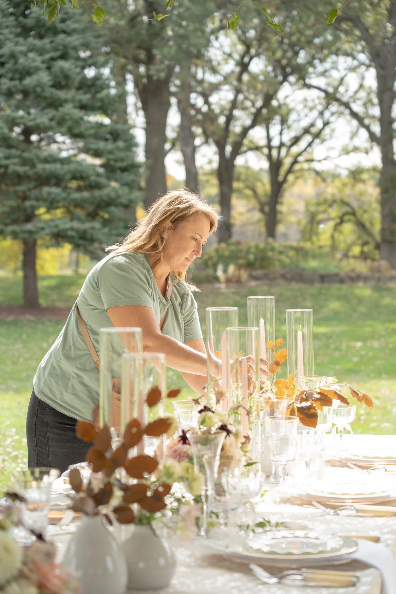 A woman arranging decor on a table outdoors in a park-like setting with trees in the background.