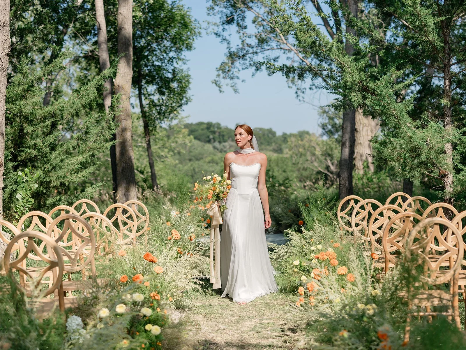 A woman in a white wedding dress standing in the middle of an outdoor garden aisle decorated with flowers, surrounded by trees and wooden chairs.