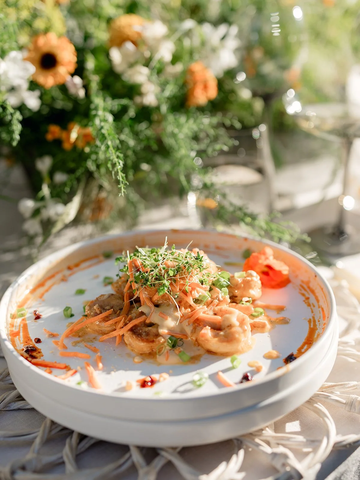 Close-up of a dish with shrimp, shredded carrots, green onions, sprouts, and a creamy sauce, served on a white plate outdoors with a blurred background of flowers and greenery.