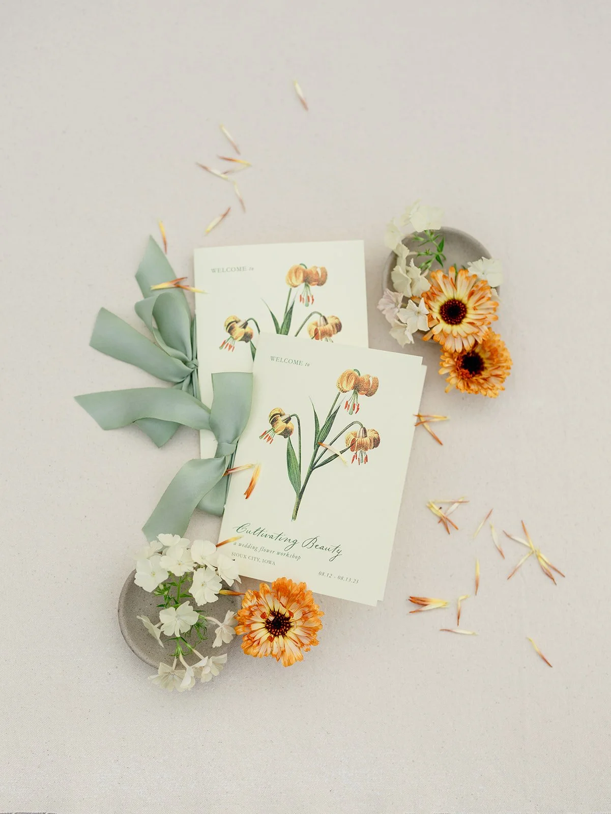 A flat lay of two wedding invitations with floral illustrations, surrounded by white and orange flowers and fallen flower petals on a white background.