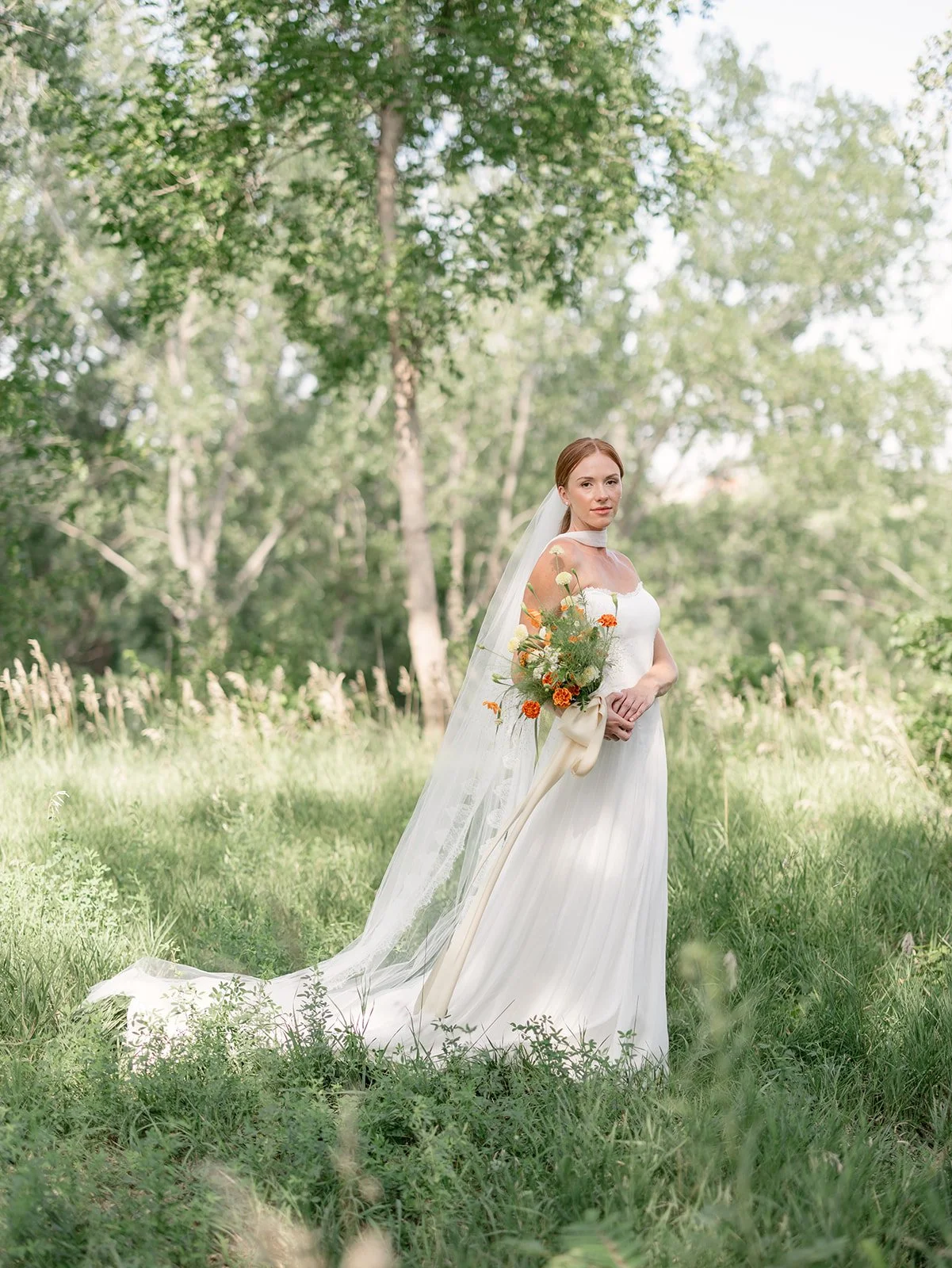 A bride in a white wedding dress holding a bouquet of orange, white, and green flowers, standing in a lush green forest with tall trees and grass.