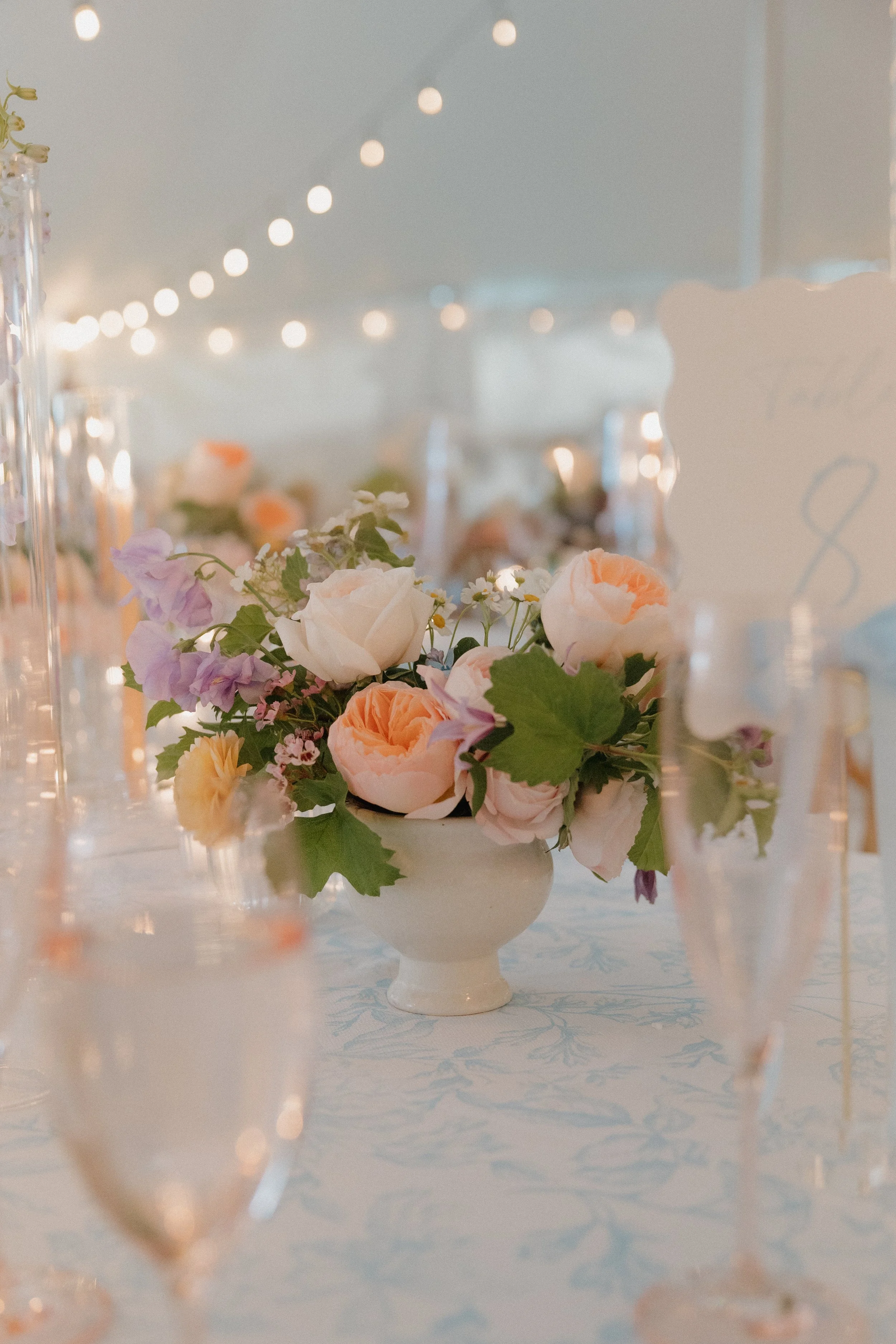 A floral centerpiece with white, peach, and purple flowers in a white ceramic vase on a table with fairy lights overhead.