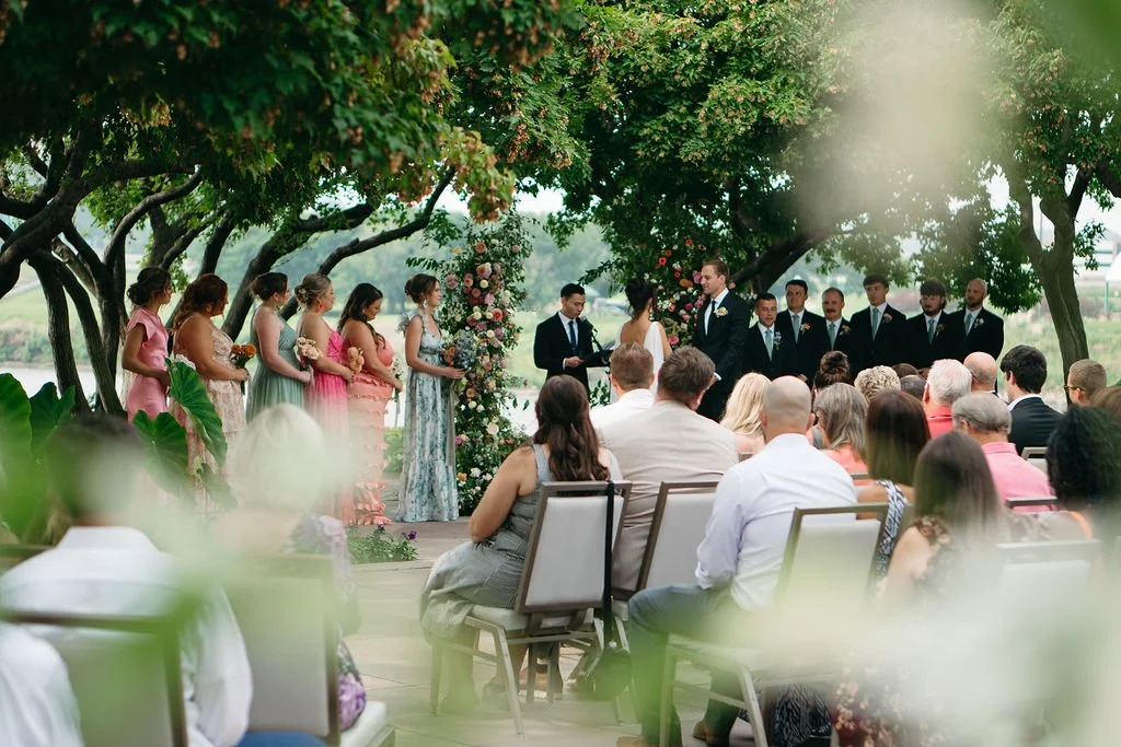 Outdoor wedding ceremony with bride and groom exchanging vows under a floral arch, surrounded by bridesmaids and groomsmen, with guests seated and watching, amid lush green trees.