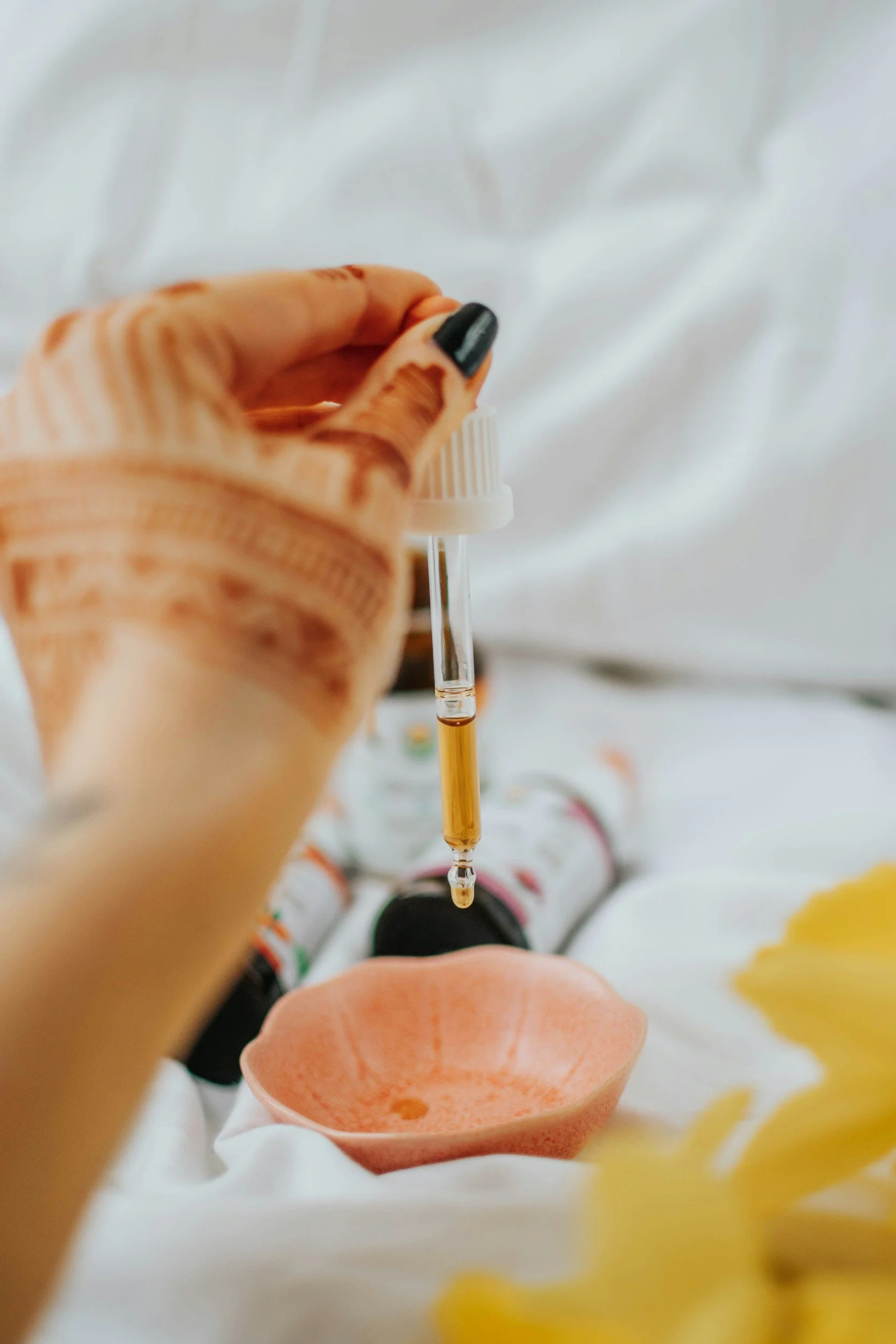 A hand holding a dropper with liquid above a bowl, surrounded by bottles and a flower, with henna on the hand.