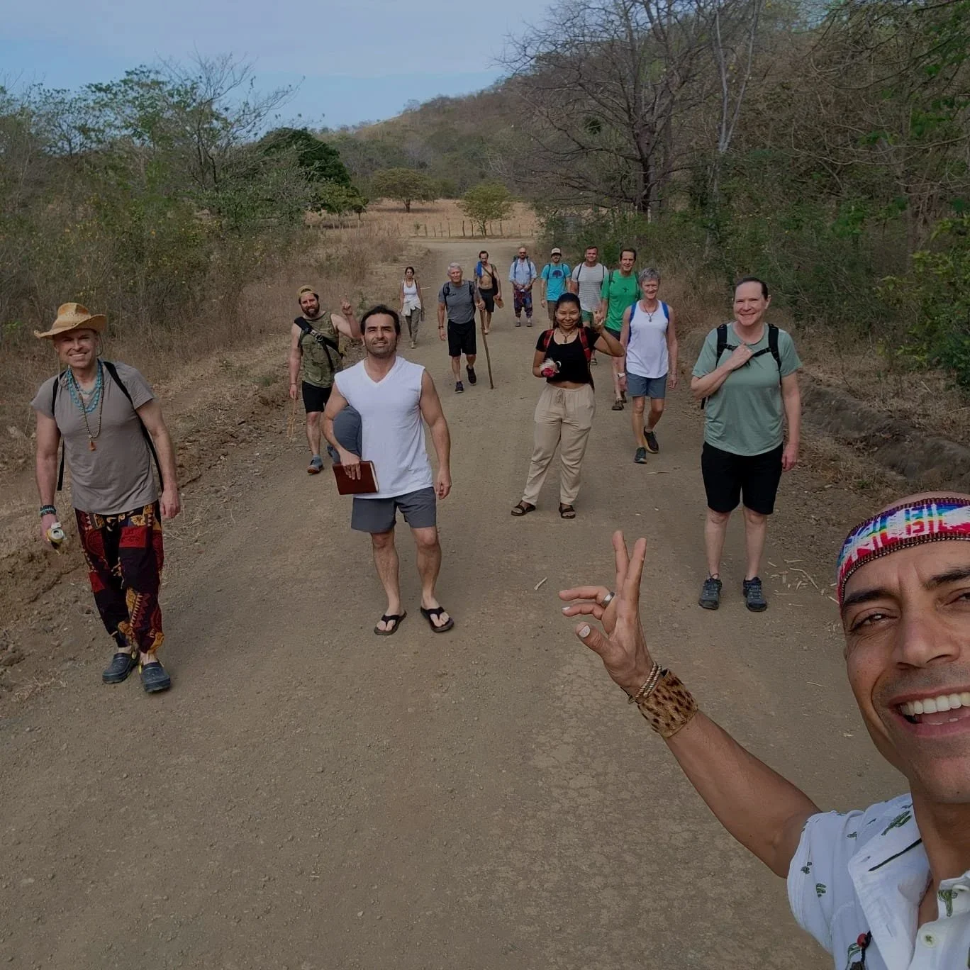 A group of people on a dirt path outdoors, some waving, smiling, and carrying backpacks, with trees and hills in the background. One person in the foreground is taking a selfie.