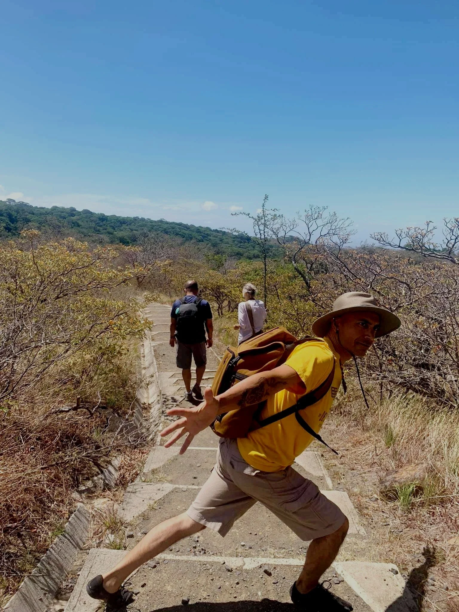 Three people hiking outdoors on a paved trail with dry grass and bushes, one man in a yellow shirt looking at camera, two others walking ahead with backpacks, hills in the background, clear blue sky.