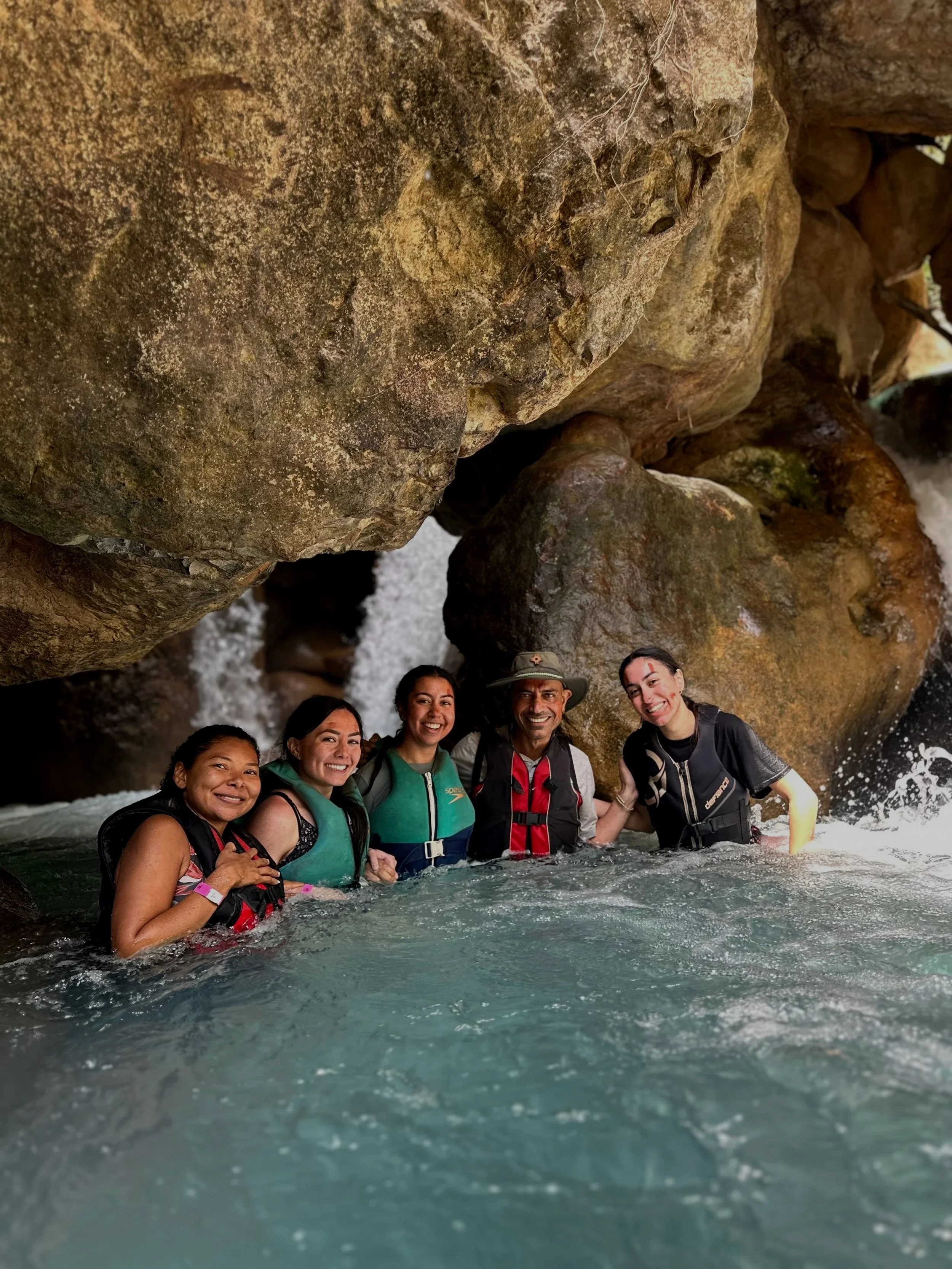 Group of five people in life jackets swimming in a river shaded by large rocks, smiling and enjoying outdoor adventure.