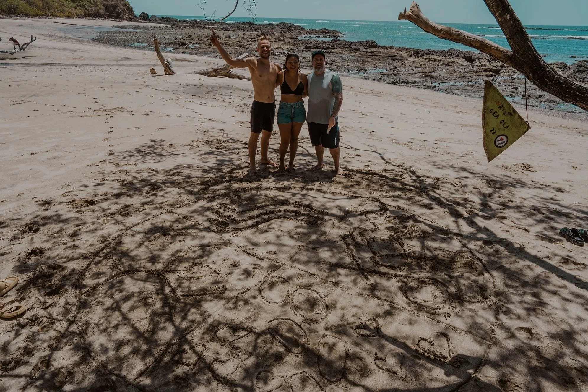 Three people standing on a beach under a tree, with a large heart and the words 'Be safe' drawn in the sand in front of them.