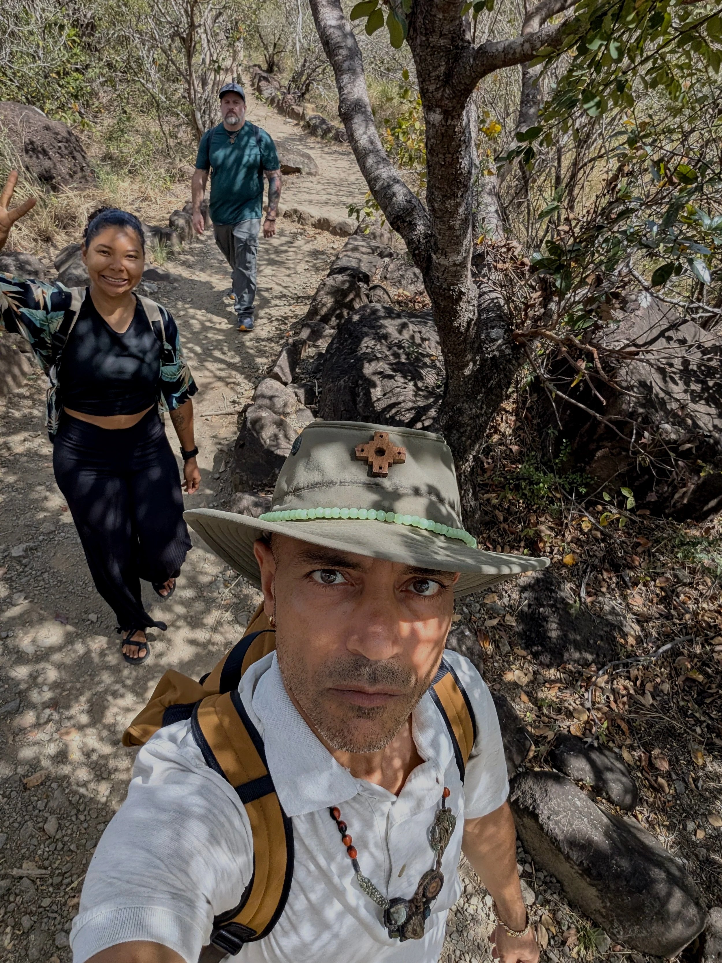 A group of three people hiking on a rocky trail in a forested area. The person in the foreground is taking a selfie, wearing a wide-brimmed hat, white shirt, and a backpack. The woman to the left is smiling and waving, dressed in black athletic cloth