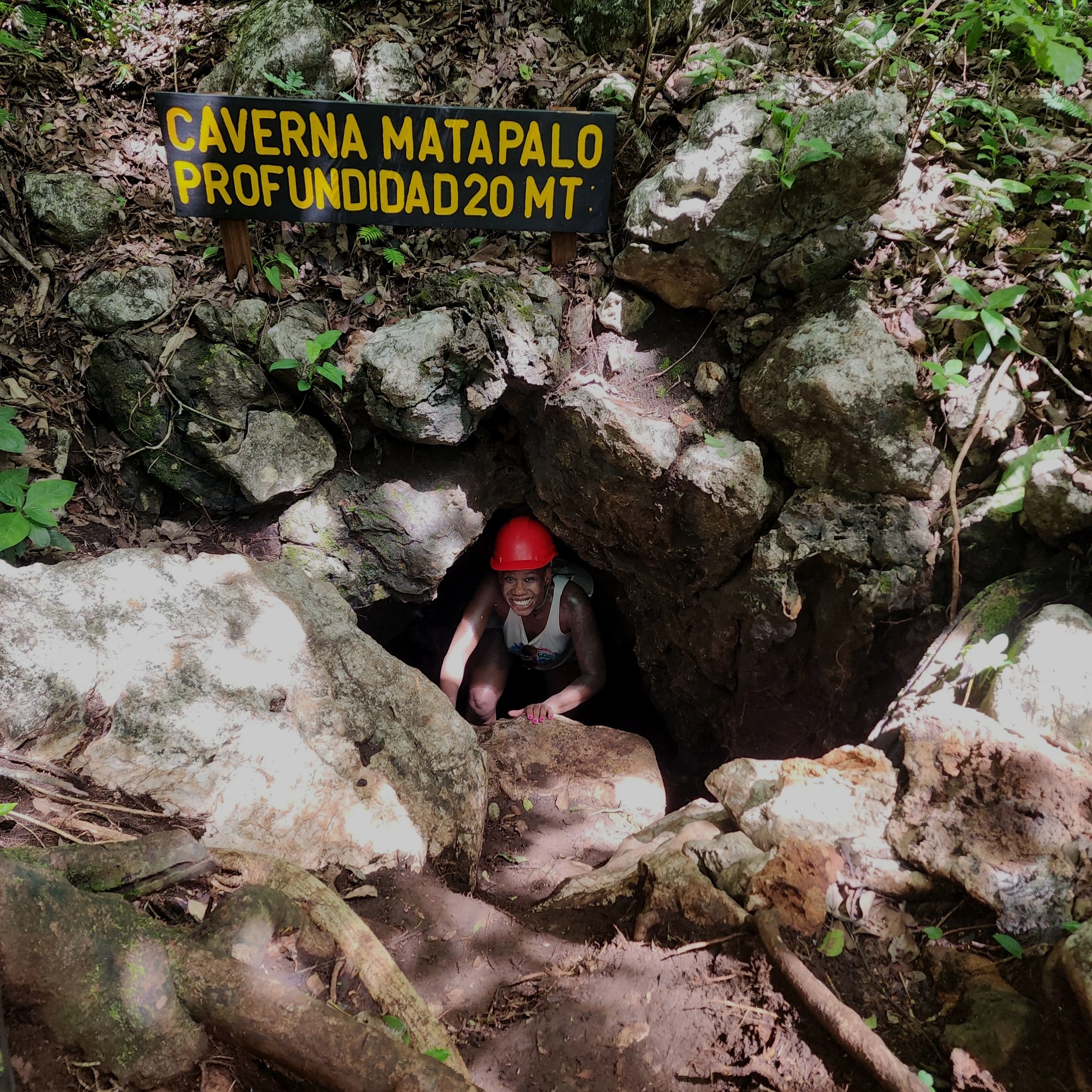 A woman wearing a red helmet crouching inside a small cave entrance surrounded by rocks and vegetation, with a sign that reads 'CAVERNA MATAPALO PROFUNDIDAD 20 MT'.