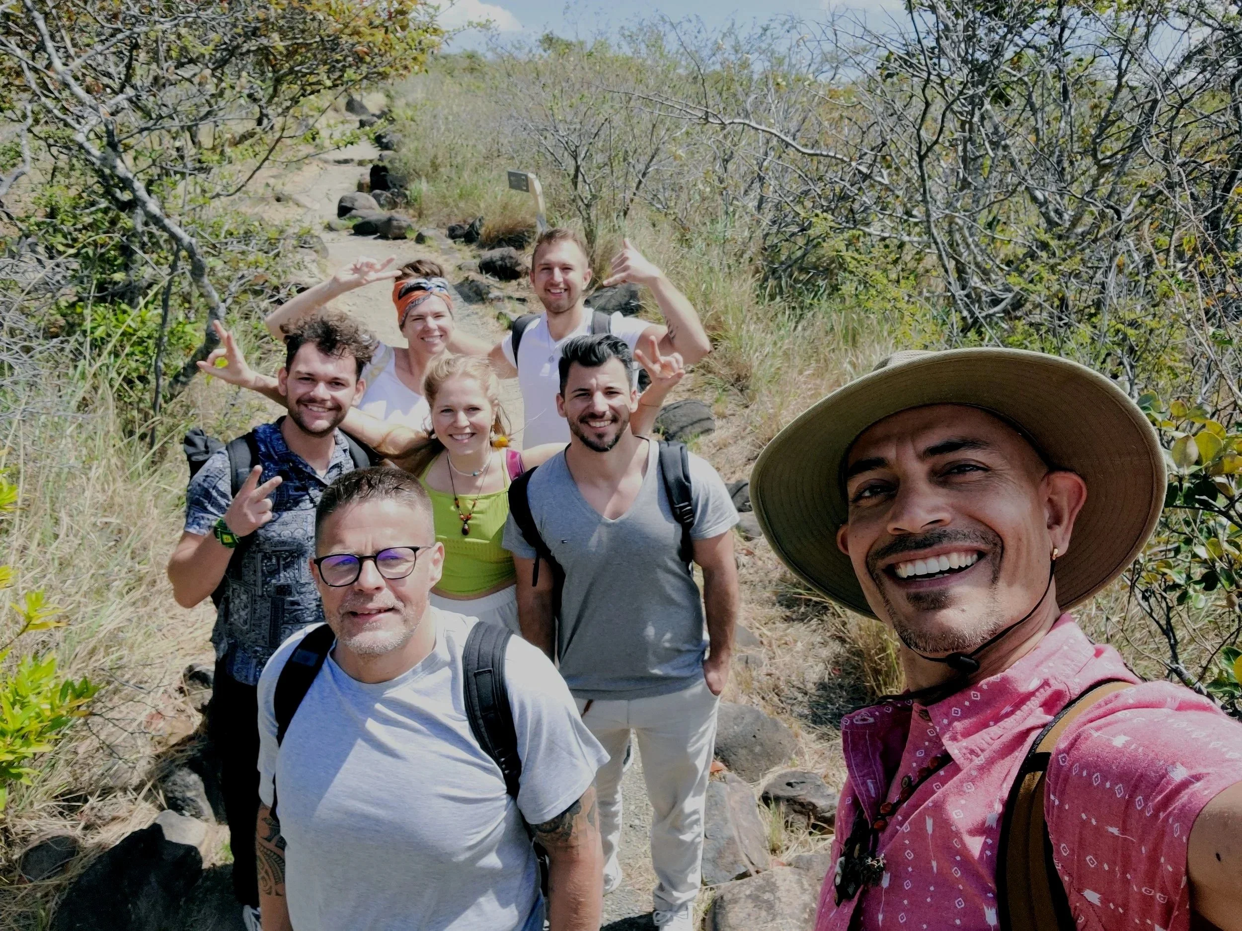 Group of eight people smiling and taking a selfie on a hiking trail surrounded by dry shrubbery and rocks.