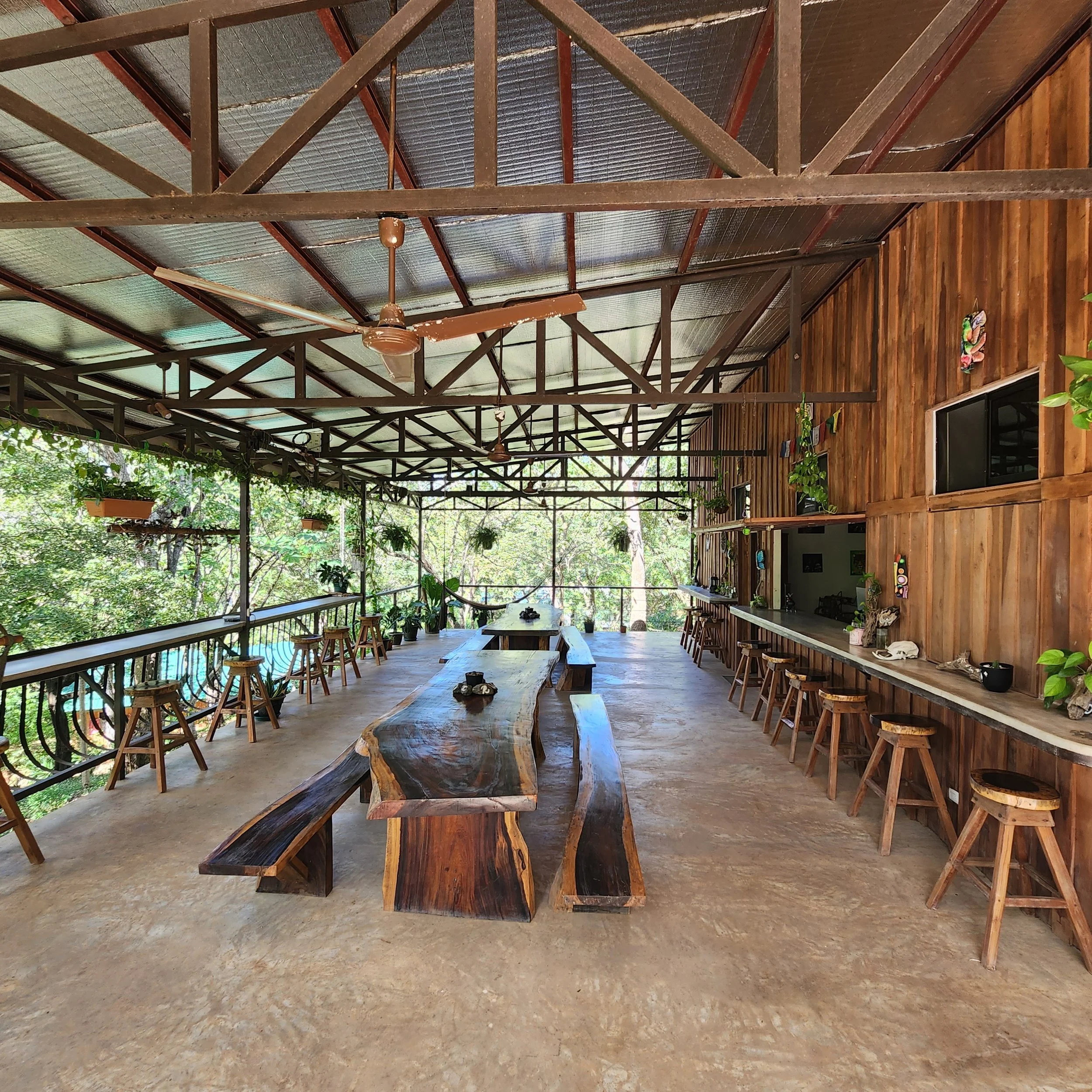 Rustic open-air cafe with long wooden tables, benches, stools, ceiling fan, plants, and a wooden backdrop.