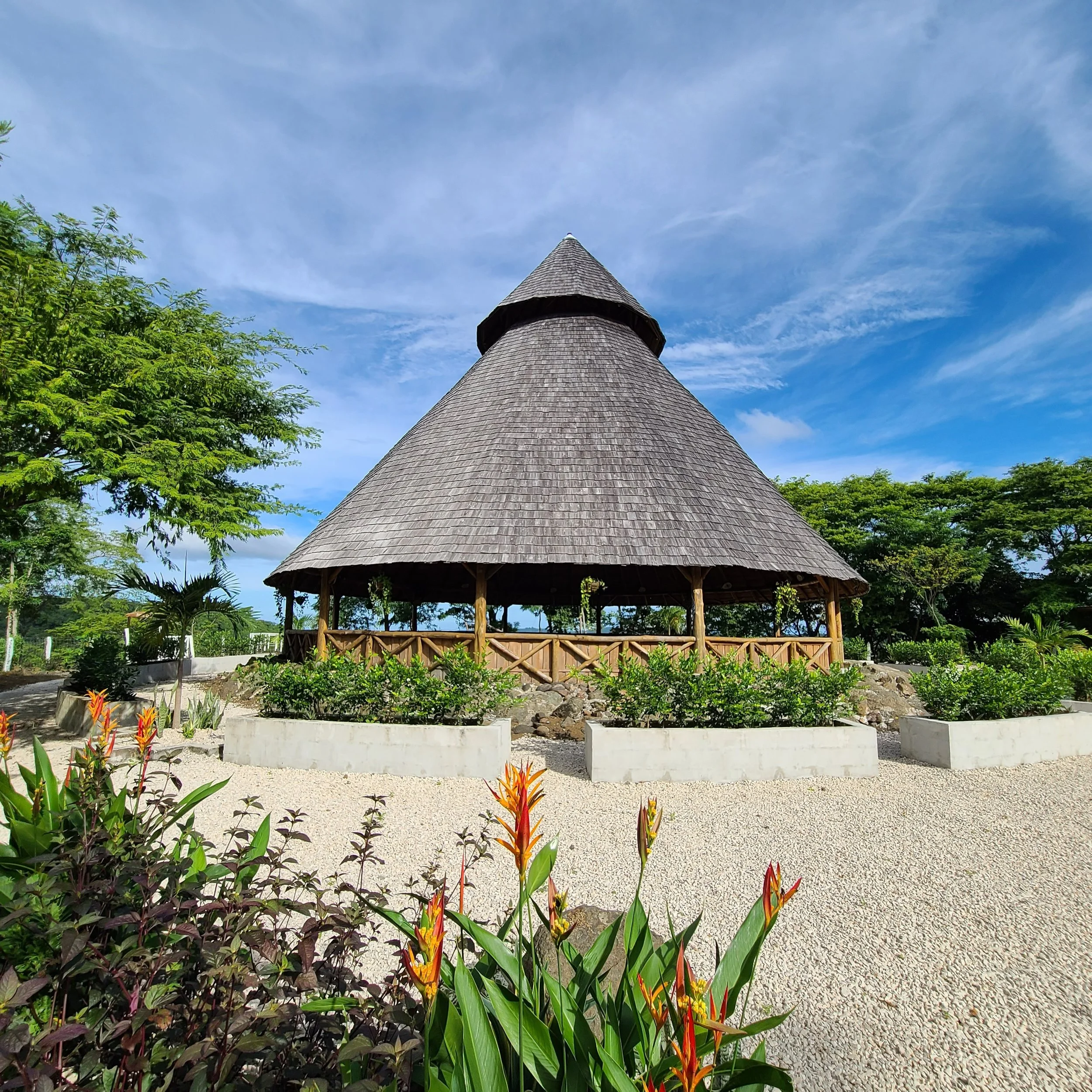 Rustic wooden pavilion with thatched roof surrounded by greenery and flowering plants under a blue sky.