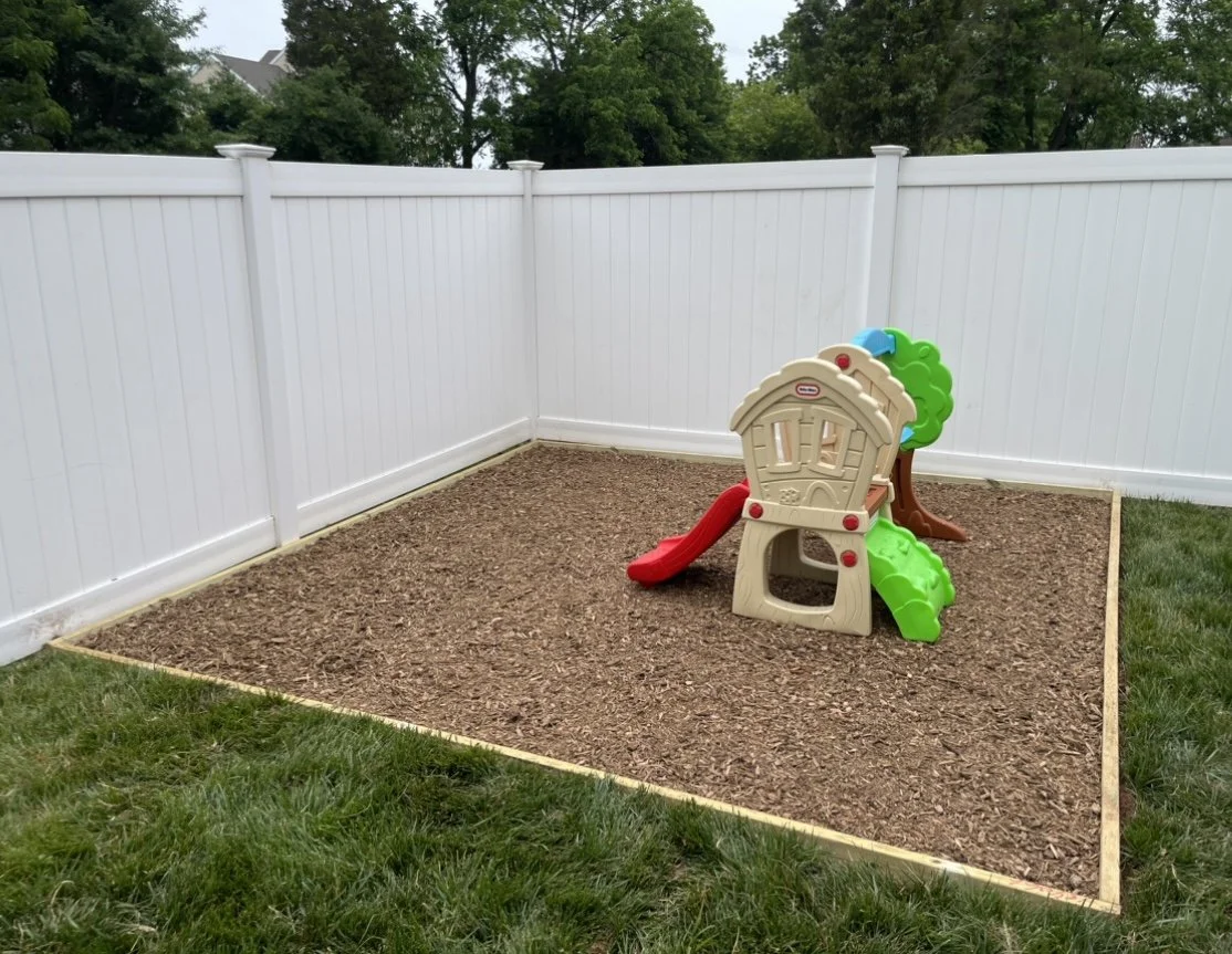Children's play area with plastic playhouse and slide on wood chips, surrounded by white fence.