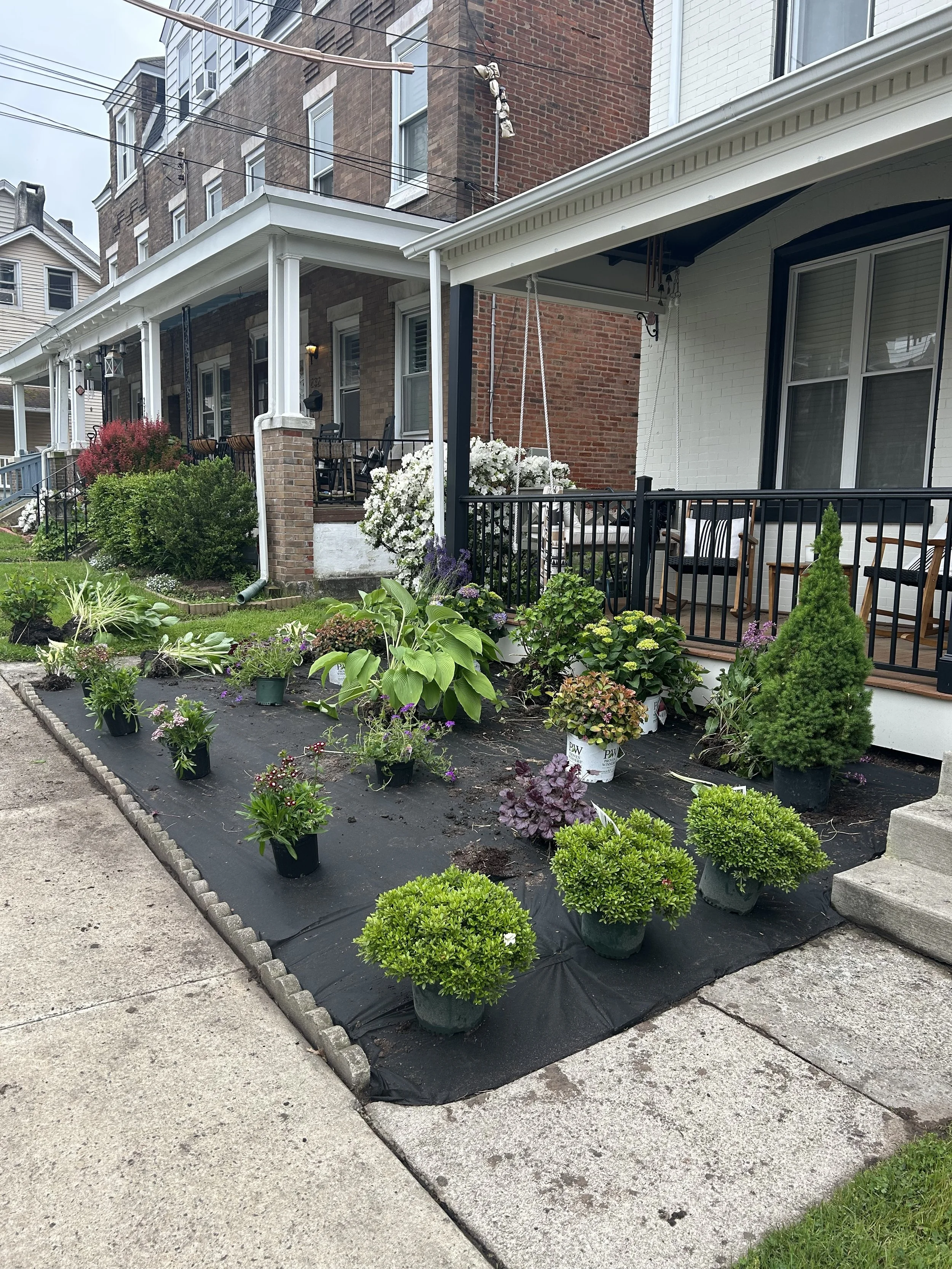 Front yard of a brick house with various potted plants on black landscaping fabric ready for planting. The house features a porch with chairs and white railing. Surrounding houses and pavement are also visible.