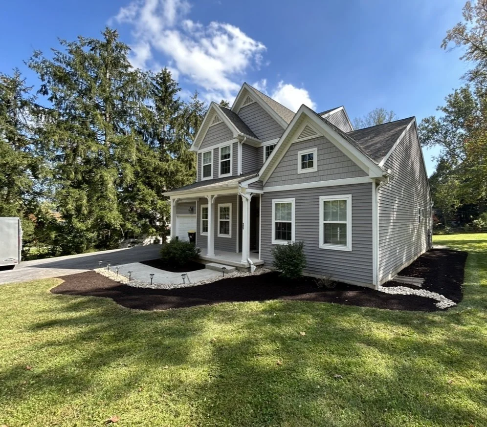 Gray two-story house with white trim, gabled roof, and driveway, surrounded by lawn and trees under a blue sky.