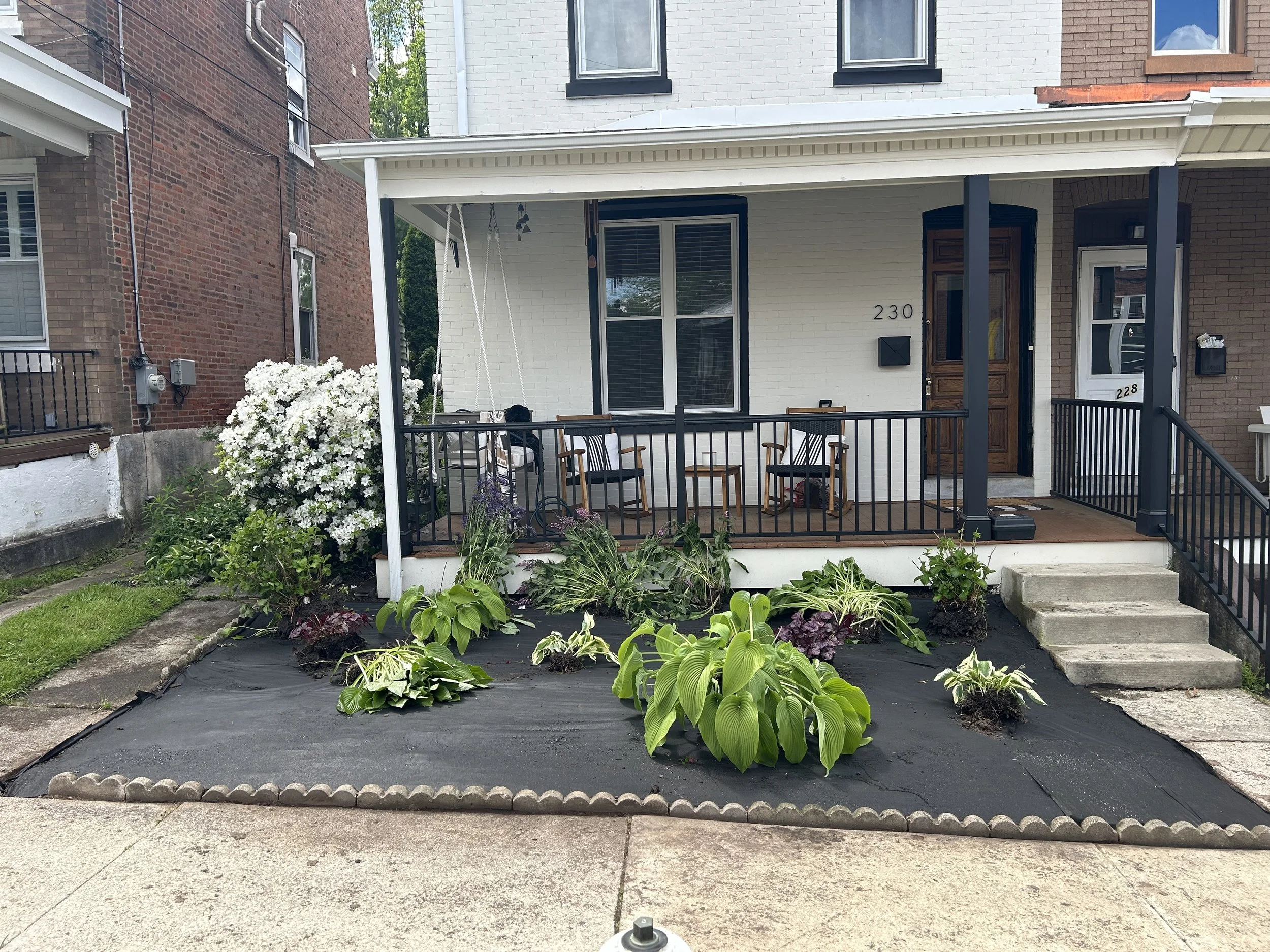 Front porch of a house with a small garden patch containing hostas and other plants, covered with black landscaping fabric. The porch has two chairs and is bordered by a black railing. A blooming bush is visible on the left side.