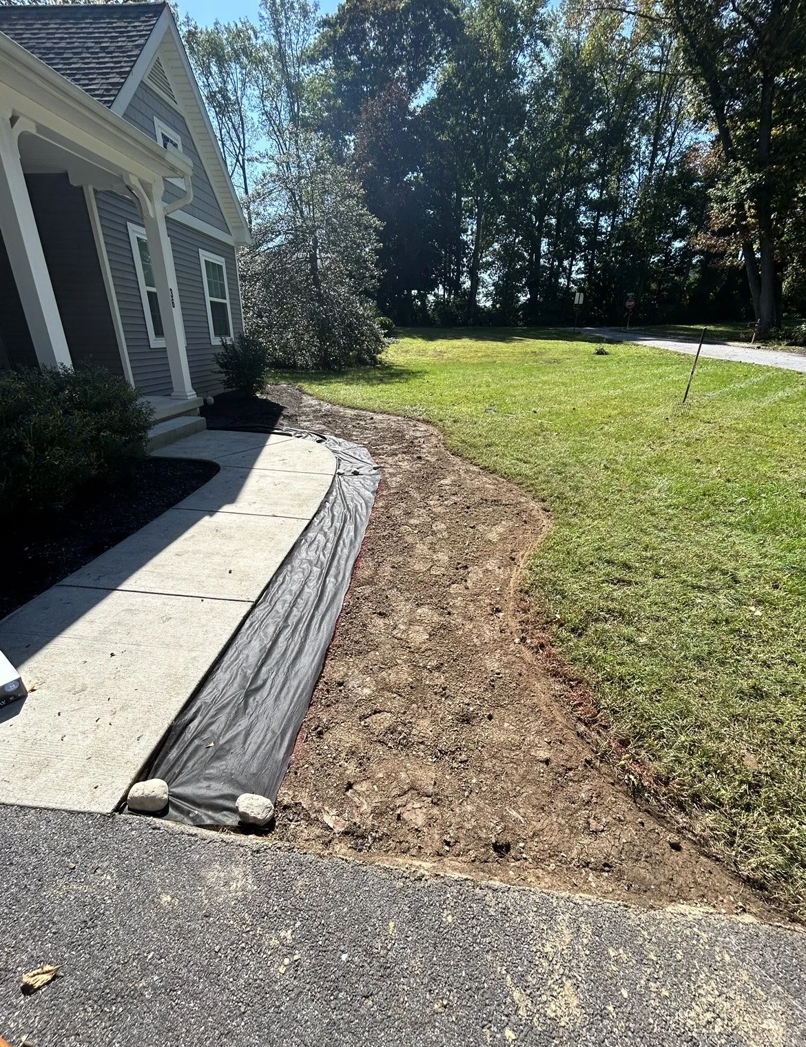 House with paved walkway, soil prepared for landscaping, grassy area, and trees in background.